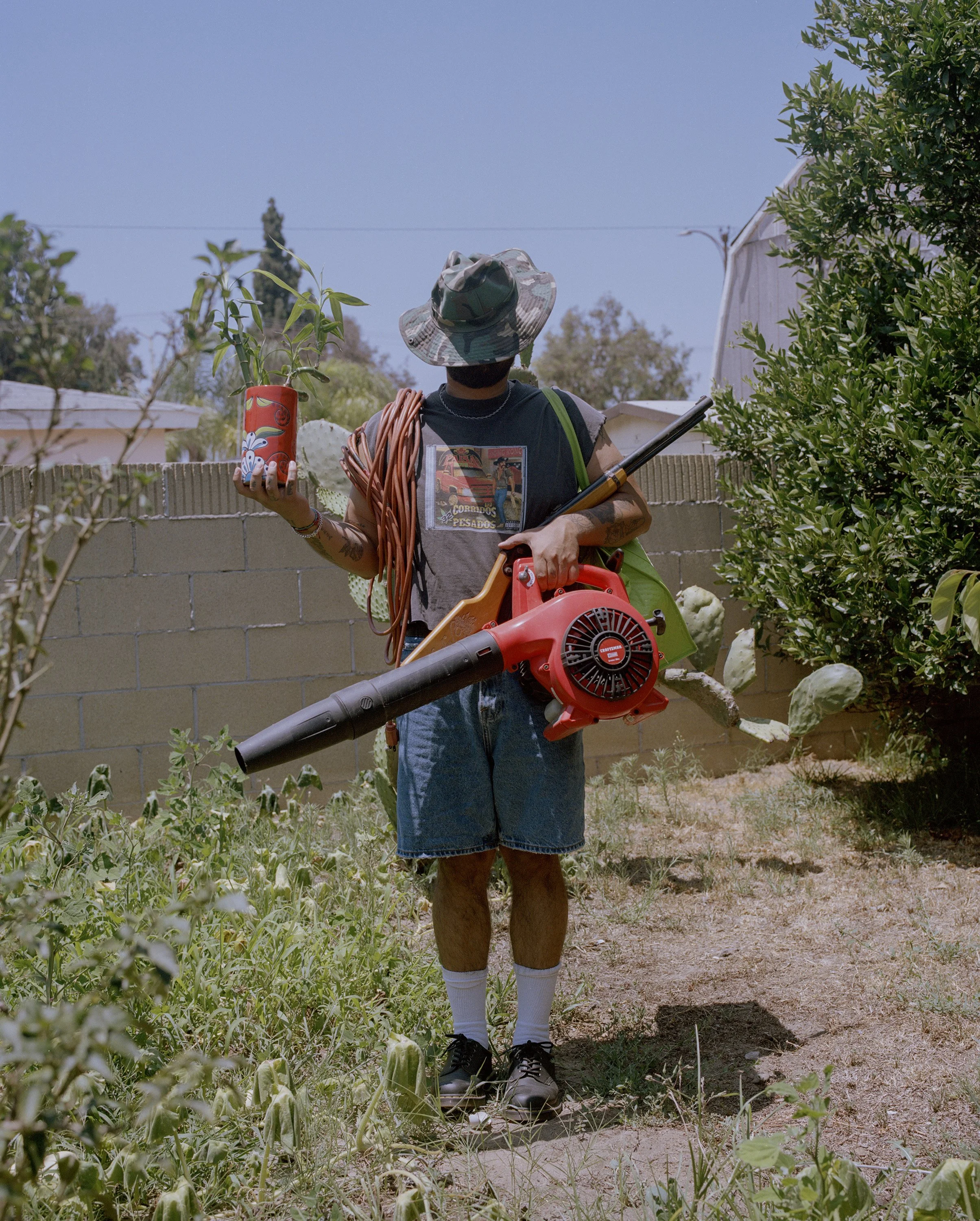 Color photograph of a man in a back yard with a hat covering his face, holding a red leaf blower, a red canister, an extension cord and a green tote bag, photo by William Camargo