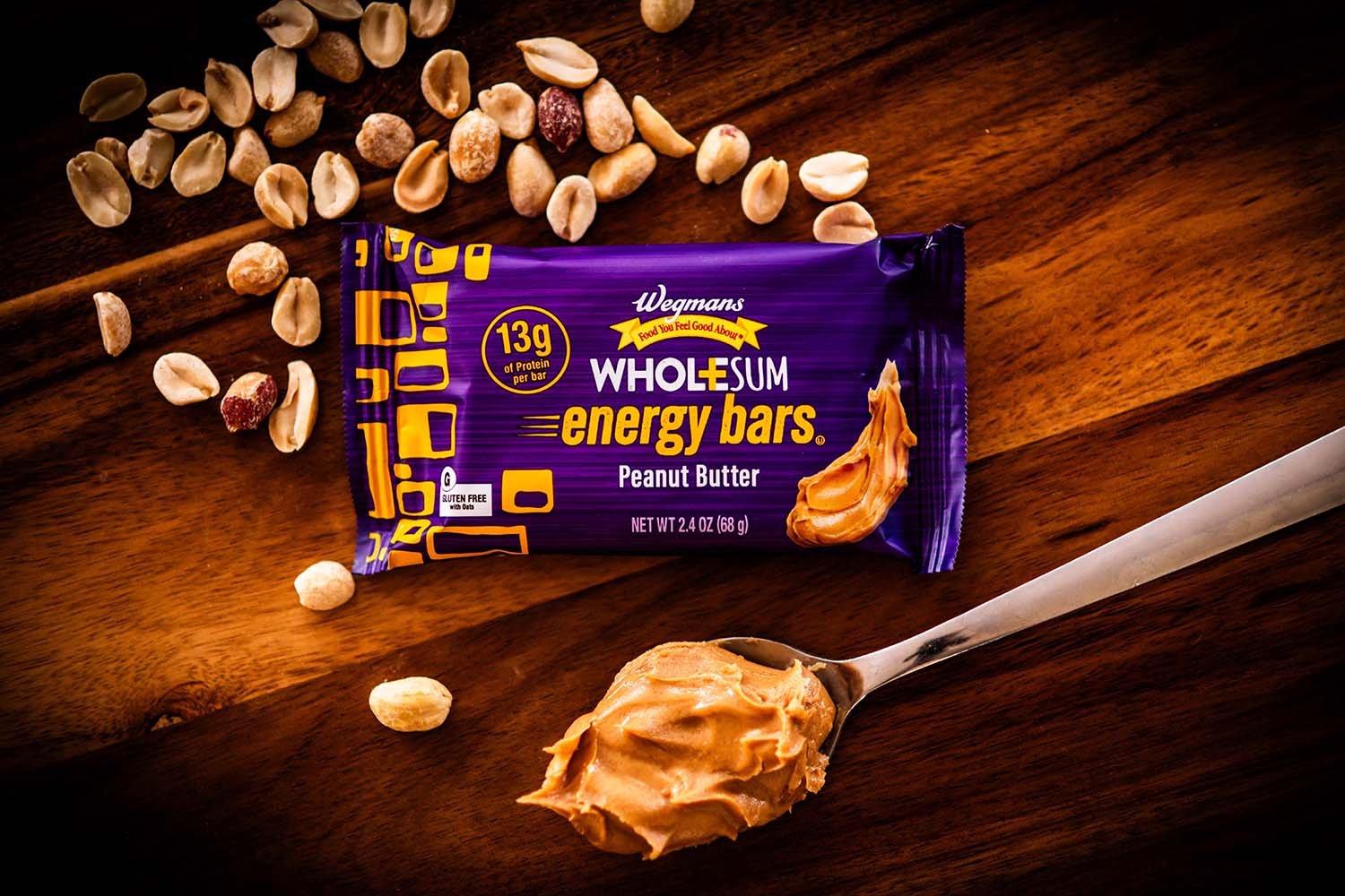 A Wegmans WholeSum peanut butter energy bar with peanut chunks around it, spilled scattered on a wooden surface, with a spoonful of peanut butter in front.