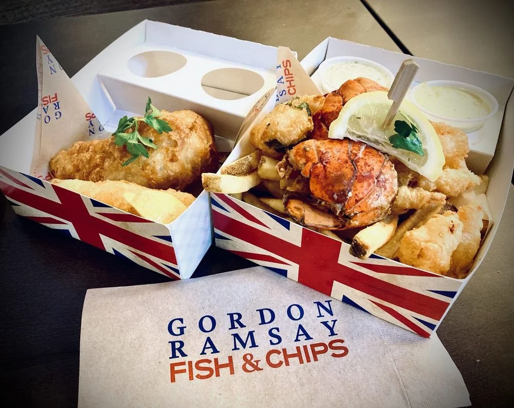 A cardboard takeout box with Union Jack design containing fried fish fillets and chips, with tartar sauce on the side. A lemon wedge with parsley is on top of the fish. There is a paper with the text 'Gordon Ramsay Fish & Chips' underneath.