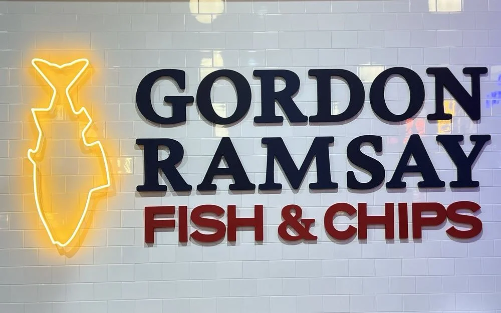 Neon sign of a fish and the text 'Gordon Ramsay Fish & Chips' on a white tiled wall.