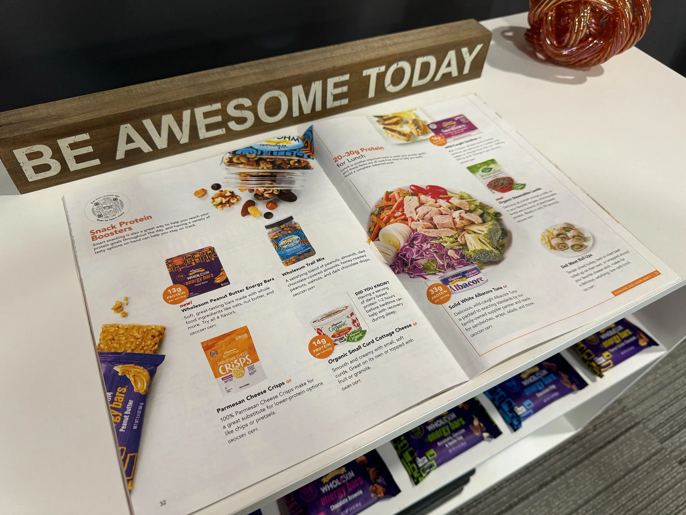Open magazine or brochure featuring a healthy food menu with snacks, salads, and protein options, placed on a white table with a wooden sign that reads "BE AWESOME TODAY" and a decorative red glass sculpture.