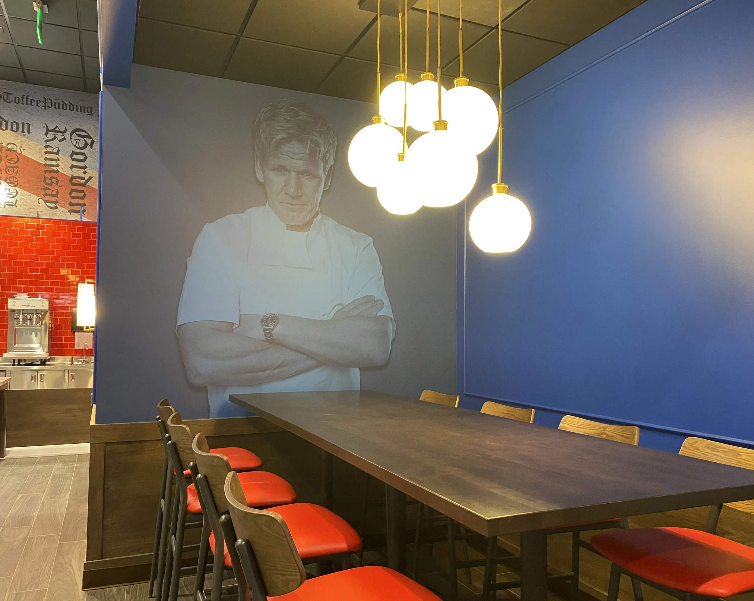 Interior of a restaurant or cafe with a large wall poster of a chef with crossed arms, white chef's coat, and a watch, hanging above a dark wooden table with red-cushioned chairs, modern hanging white globe lights, and a blue wall in the background.