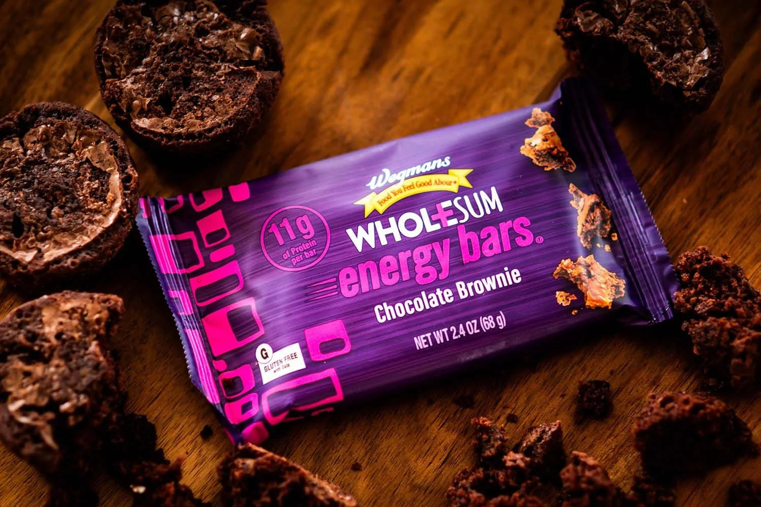 A purple package of Walgreens WholeSum energy bars, flavor Chocolate Brownie, surrounded by broken pieces of chocolate brownie on a wooden surface.