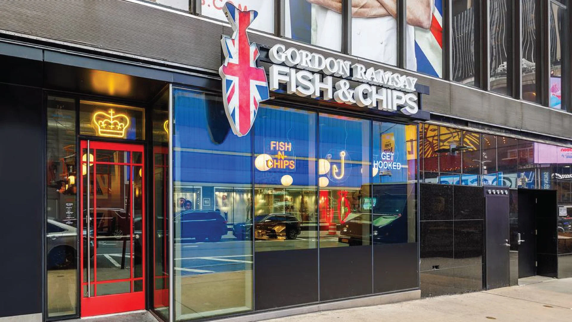 Exterior of a fish and chips restaurant with a sign that reads "Gordon Ramsay Fish & Chips" and features a Union Jack flag logo. The storefront has large reflective windows, a red door, and neon signs inside.