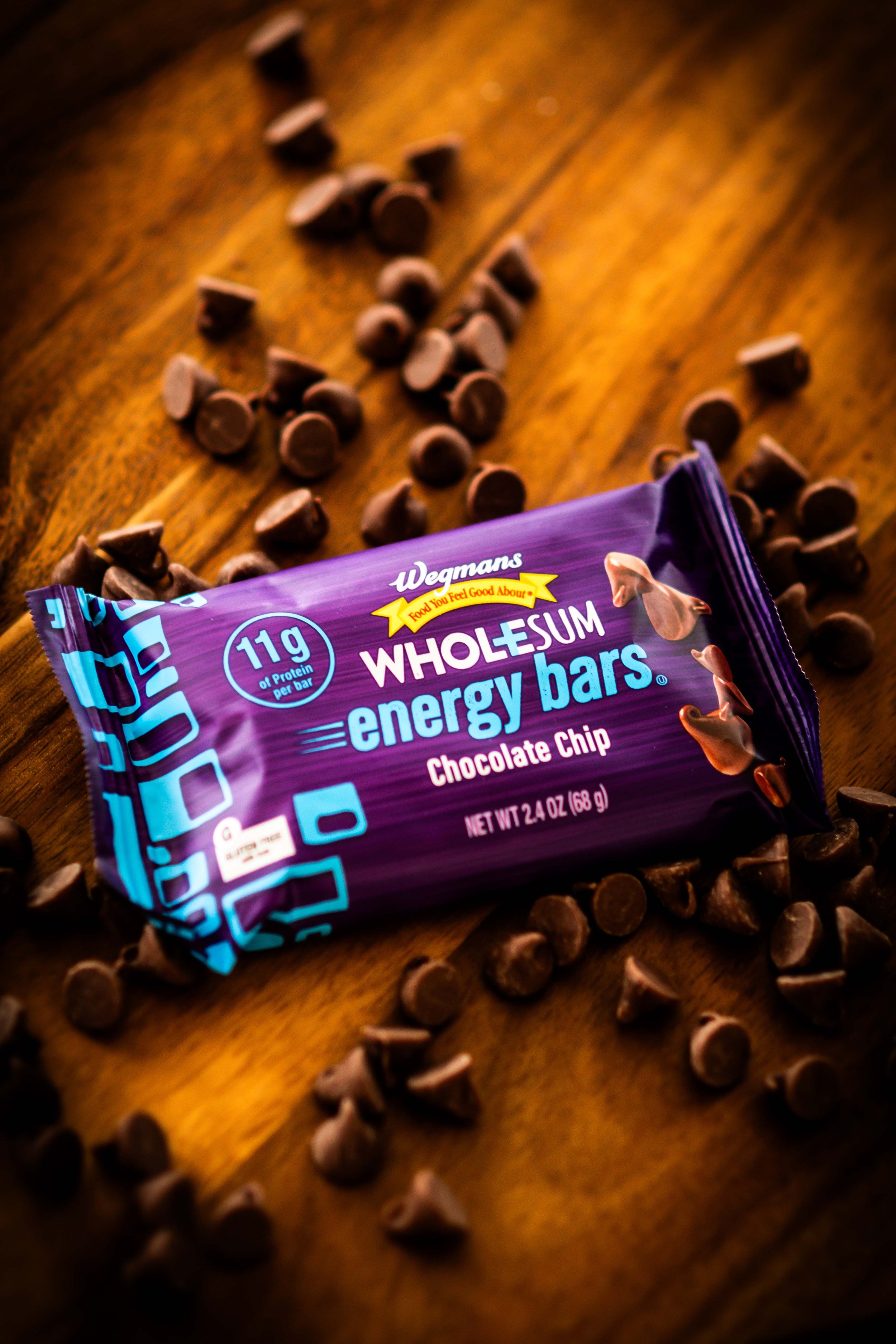 A Wegmans Whole Sum energy bar in chocolate chip flavor surrounded by scattered chocolate chips on a wooden surface.