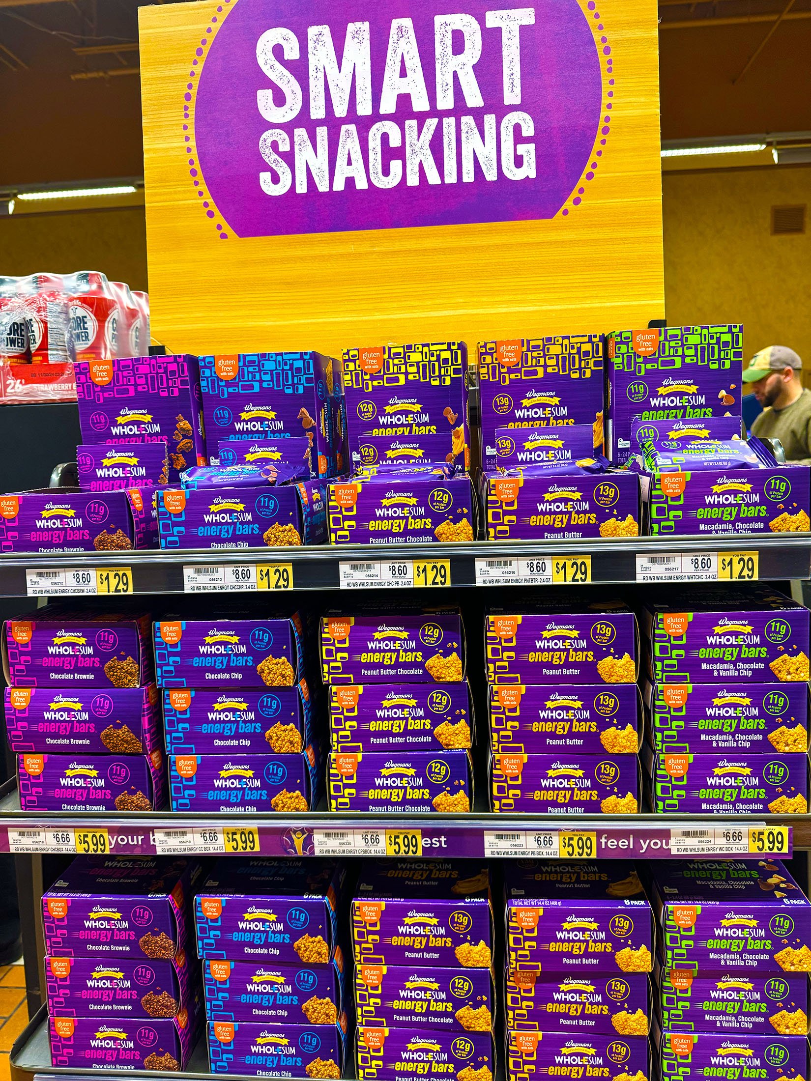Display of Wegmans energy bars in purple, blue, yellow, and green packaging with a sign that reads 'Smart Snacking' in a store aisle.