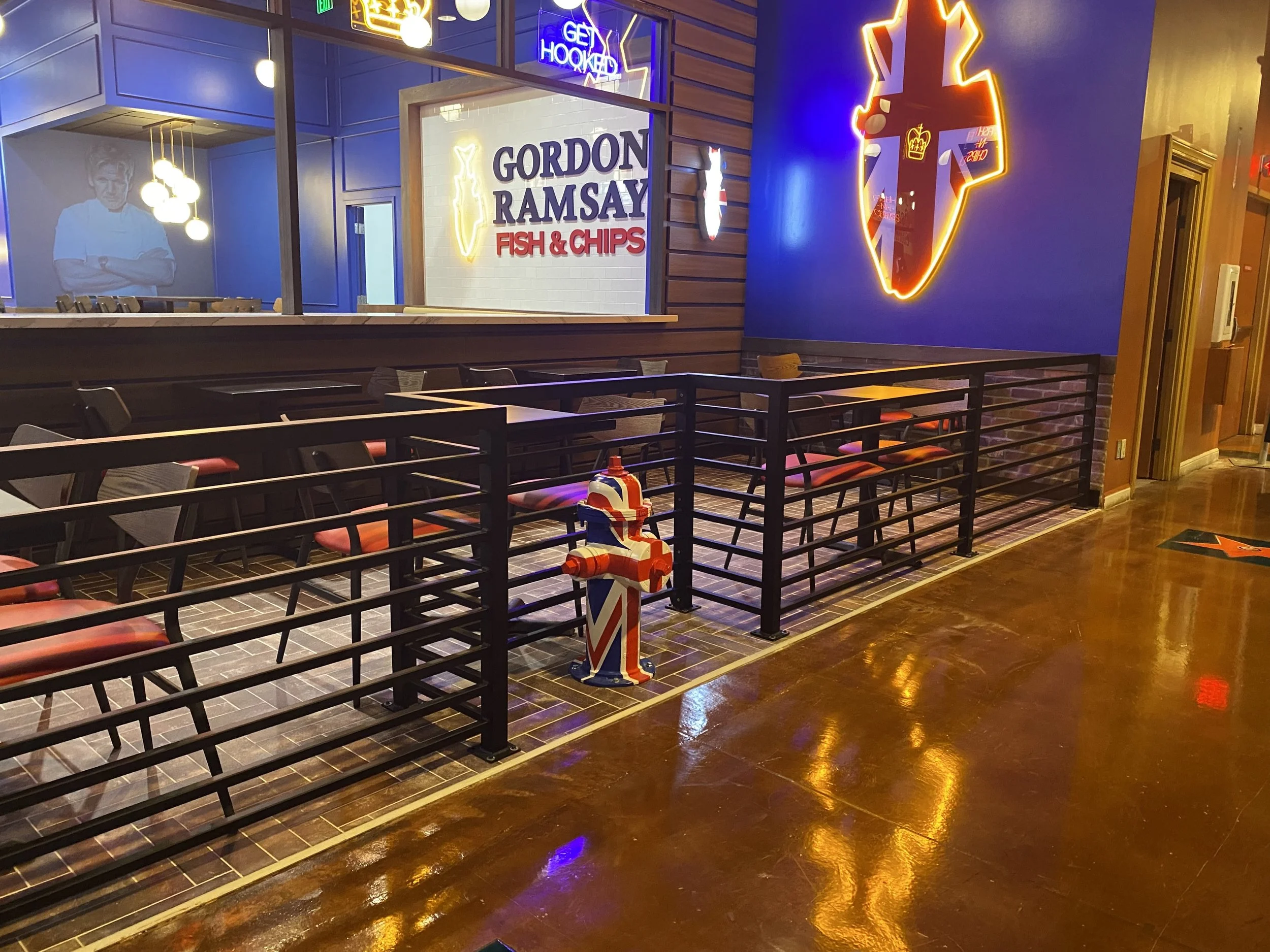 Empty seating area with black metal fence and pink cushioned chairs in a restaurant or pub with neon signs, including a blue sign that says 'GET HOOKED,' a sign for 'GORDON RAMSAY FISH & CHIPS,' up a wall, and a fire hydrant with Union Jack pattern.