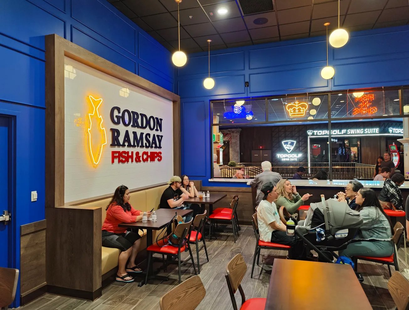People dining inside a Gordon Ramsay Fish & Chips restaurant, with neon signs and a blue wall, and other patrons visible through the window at Topgolf Swing Suite.