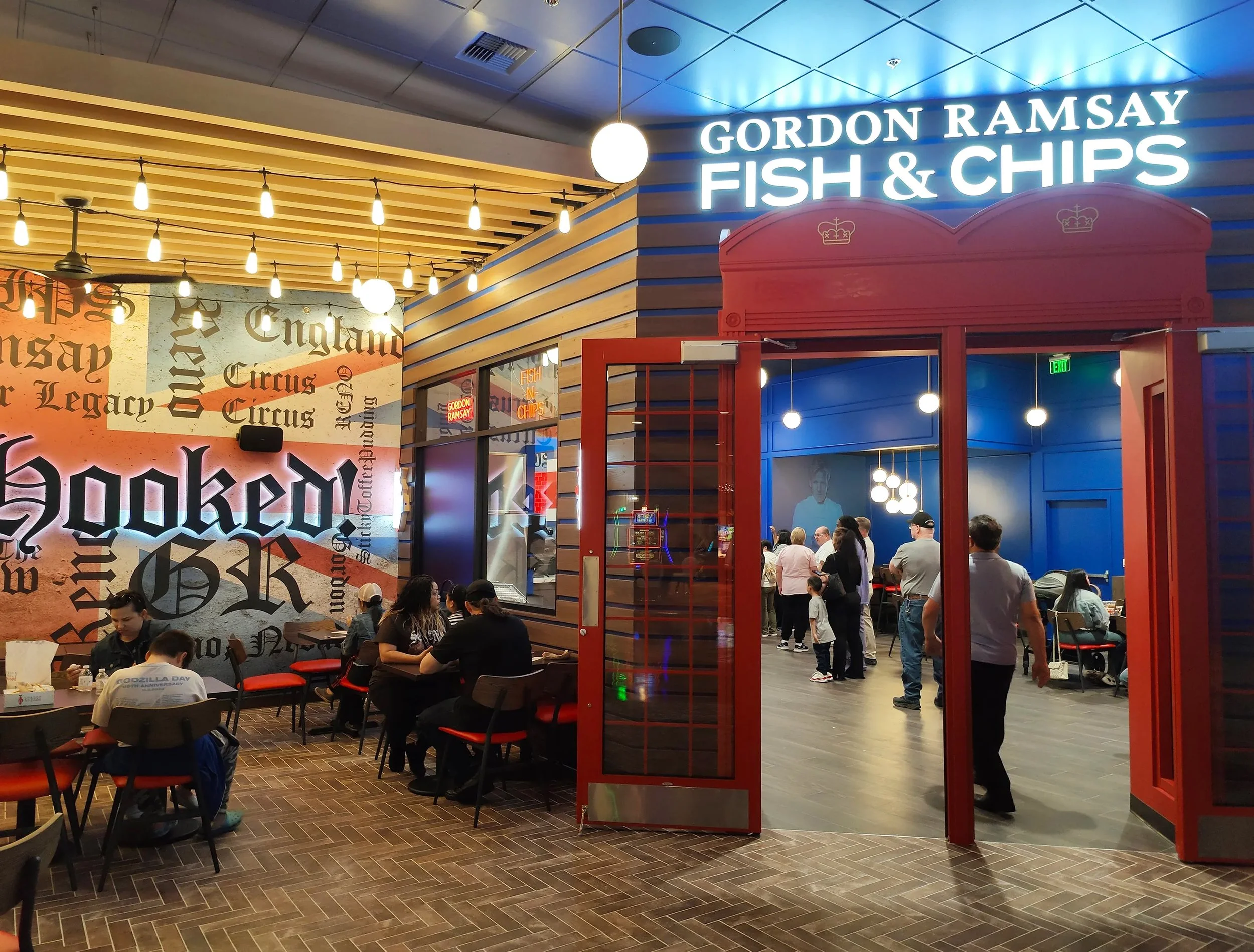 Inside Gordon Ramsay Fish & Chips restaurant, with groups of people dining and standing in line, illuminated signage, and decorative wall art.