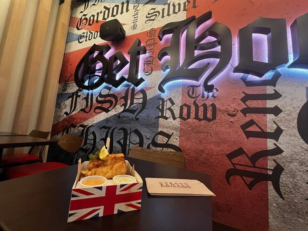 A table with a cardboard food container featuring the Union Jack flag, containing fried fish, lemon, and sauces, in a British-themed restaurant with a graffiti-style wall that says 'Get Ripped' and other words like 'Hush' and 'Row'.