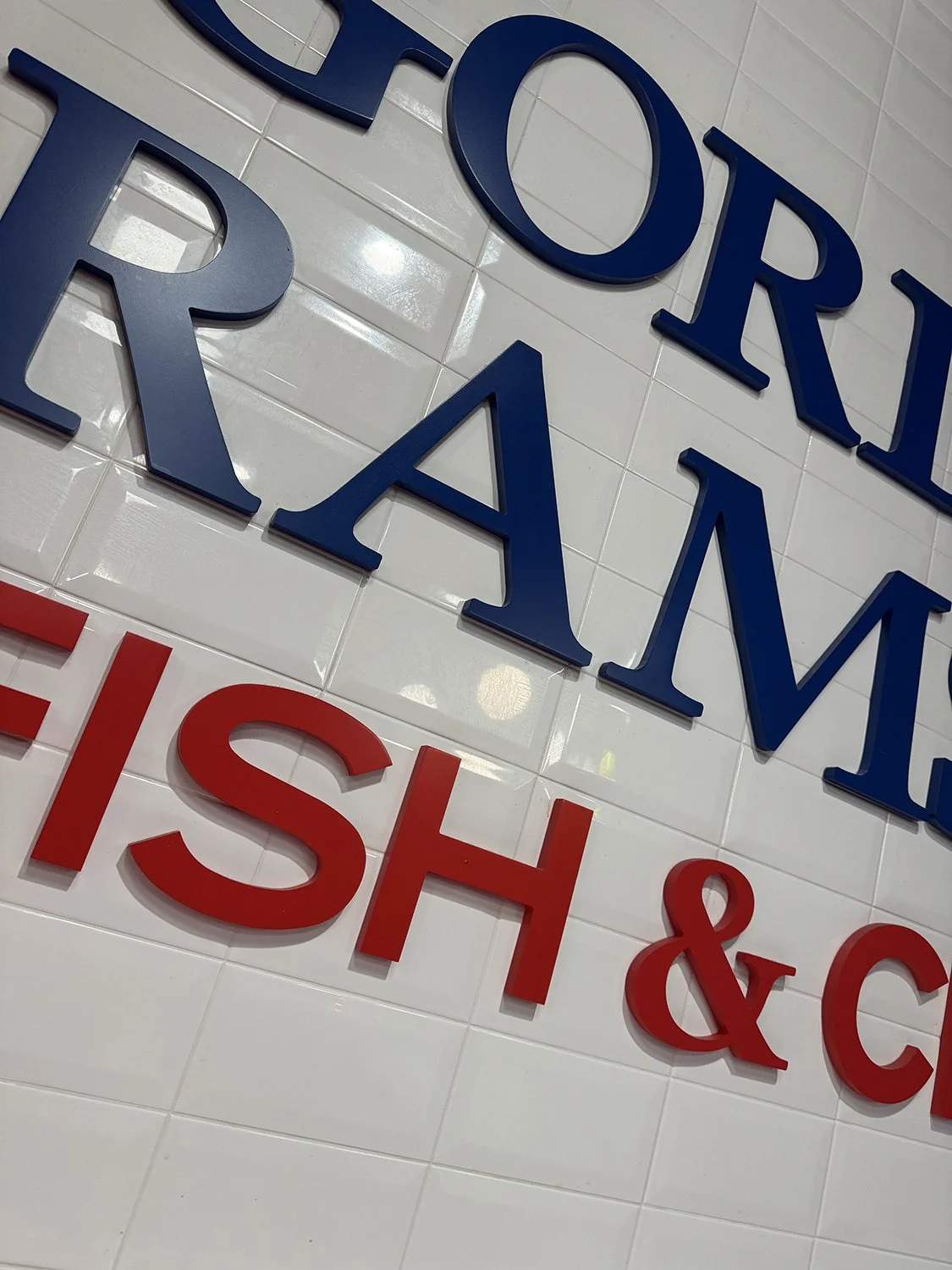 Close-up of a wall with blue and red cut-out letters spelling 'GORDON RAMSAY FISH & CHIPS'. The wall is tiled with white glossy tiles.