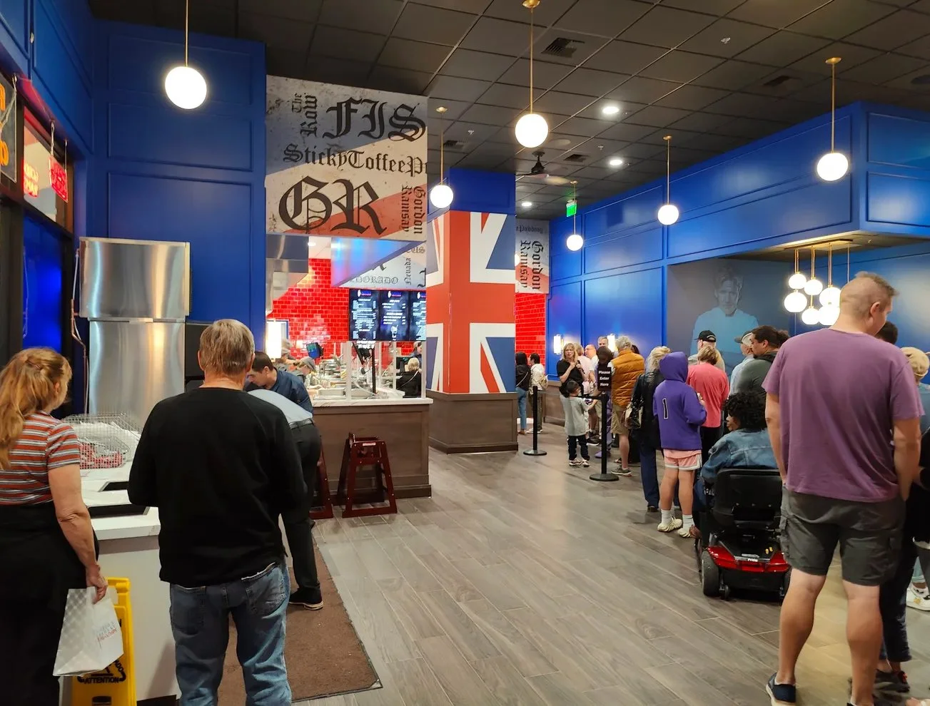 Inside a busy cafe with blue walls, large British flag decoration, and hanging globe lights, people are lined up waiting to order or sitting at tables.