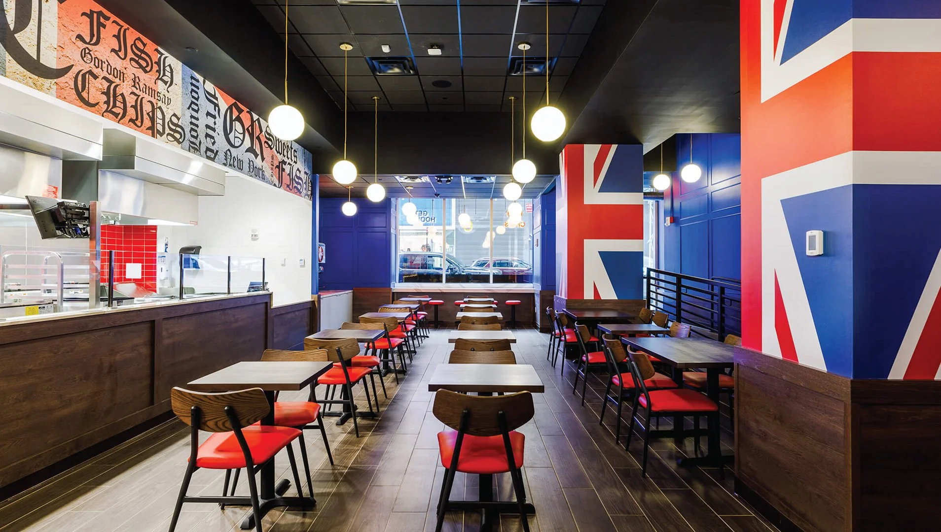 Empty restaurant decorated with British flag motifs, featuring wooden tables and chairs with red seats, large windows at the back, and hanging round lights.