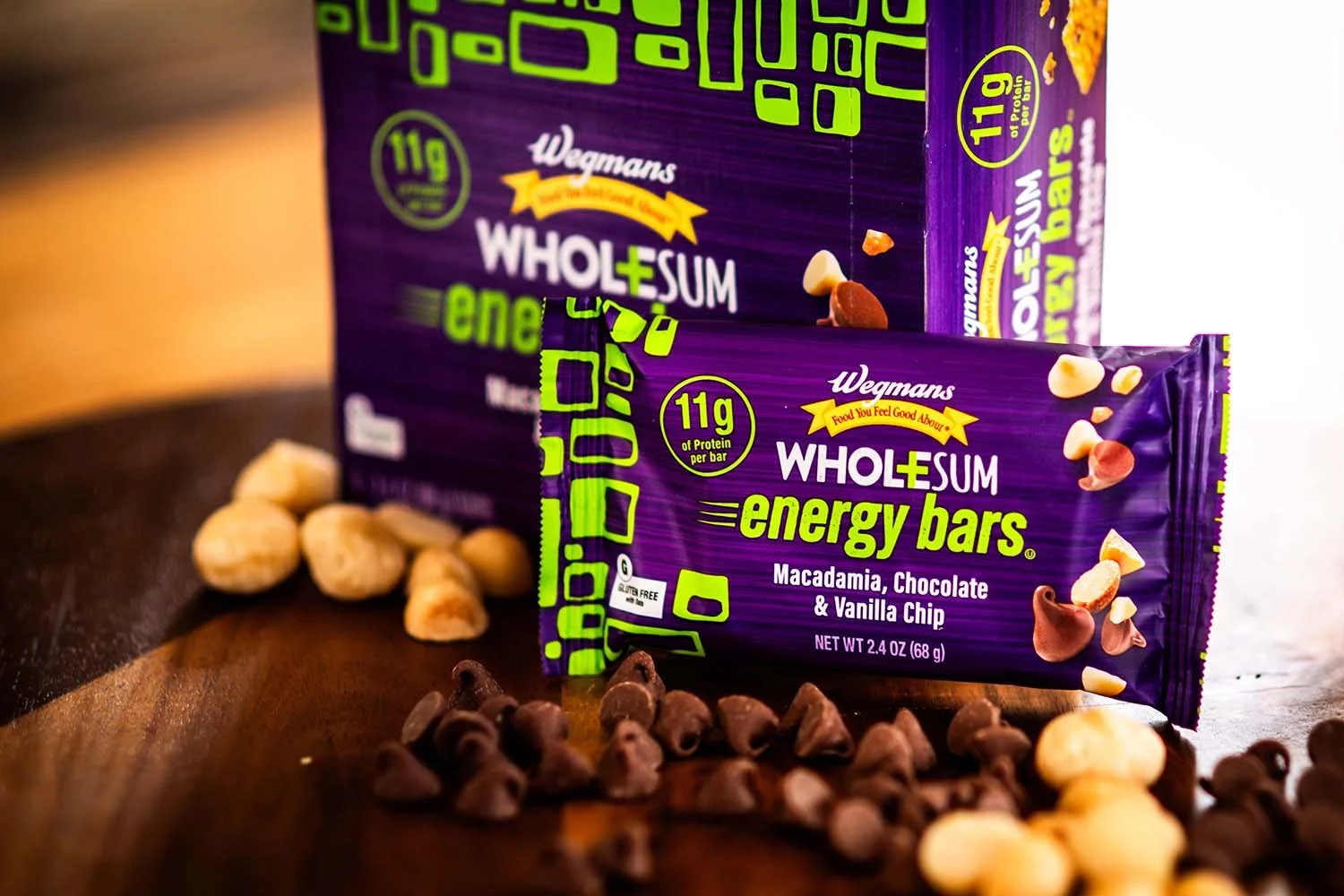 A purple box of Wegmans WholeSum energy bars with chocolate, vanilla, and macadamia flavor on a wooden surface with scattered white and dark chocolate chips.