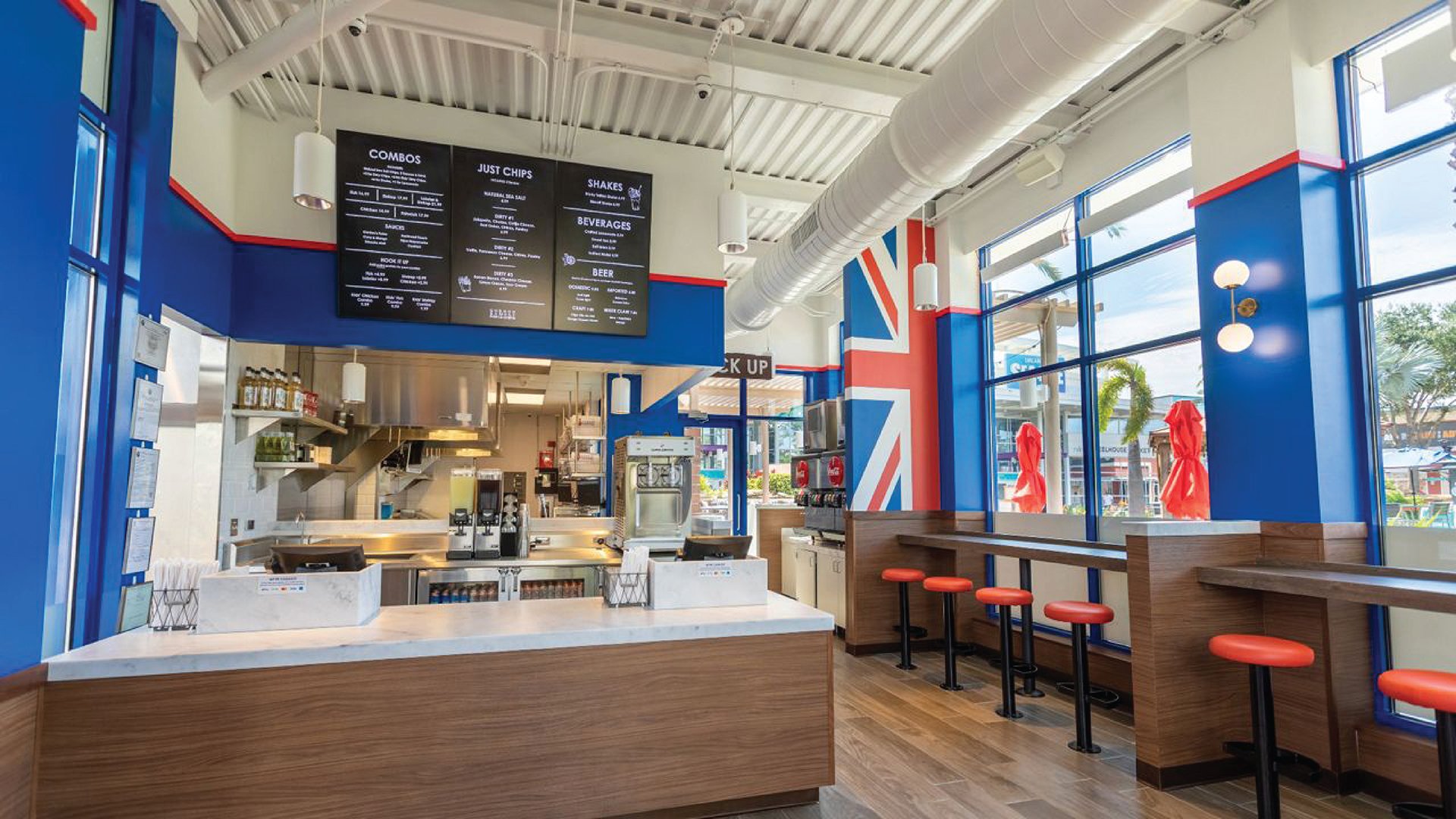 Interior of a brightly lit fast food restaurant with blue walls and a British flag mural, seating with red stools at a high table, and an order counter with a menu board overhead.