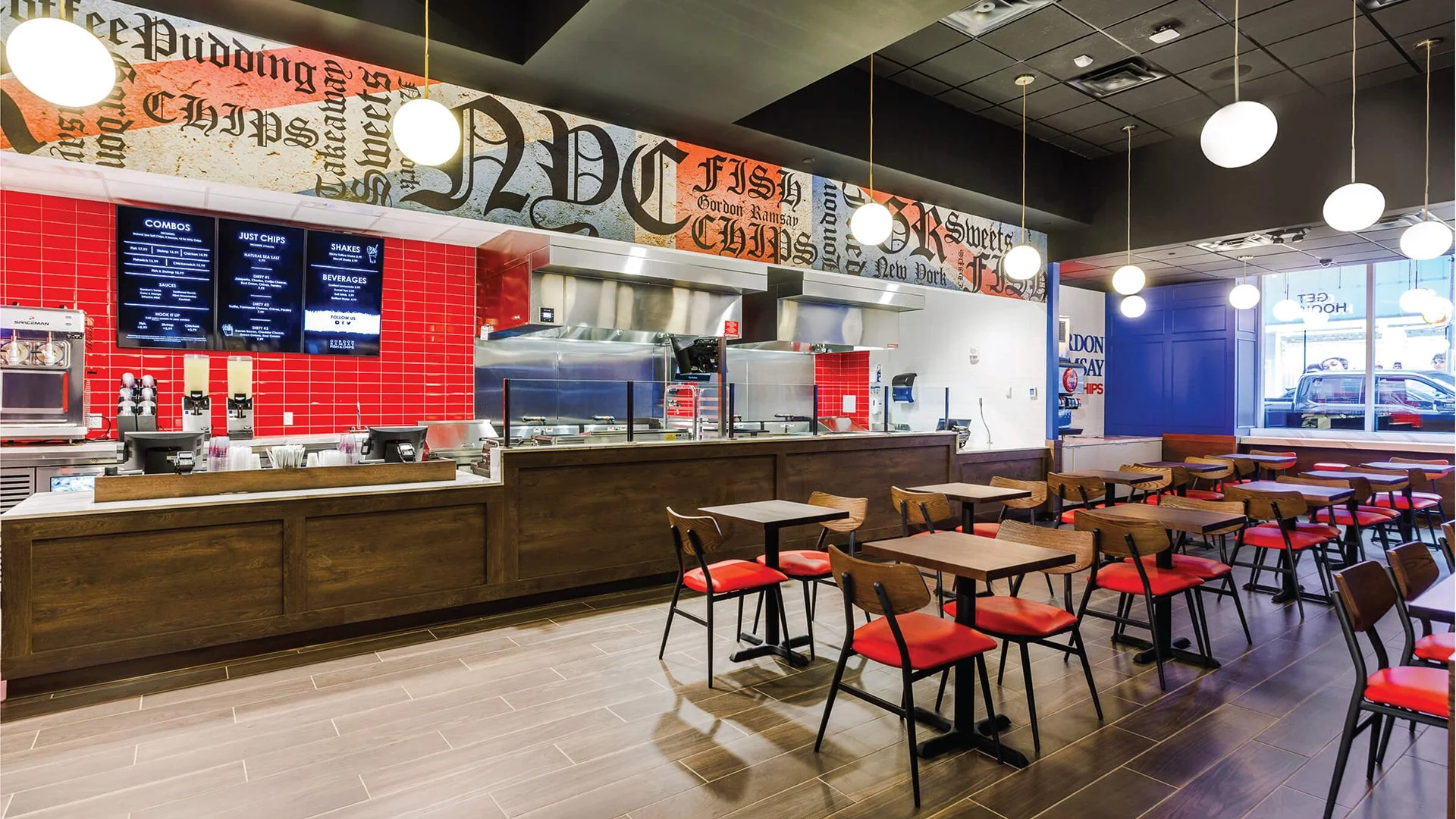 Interior of a modern restaurant with wooden tables and red cushioned chairs, a red tile counter, digital menu screens, and a large window showing parked cars outside.