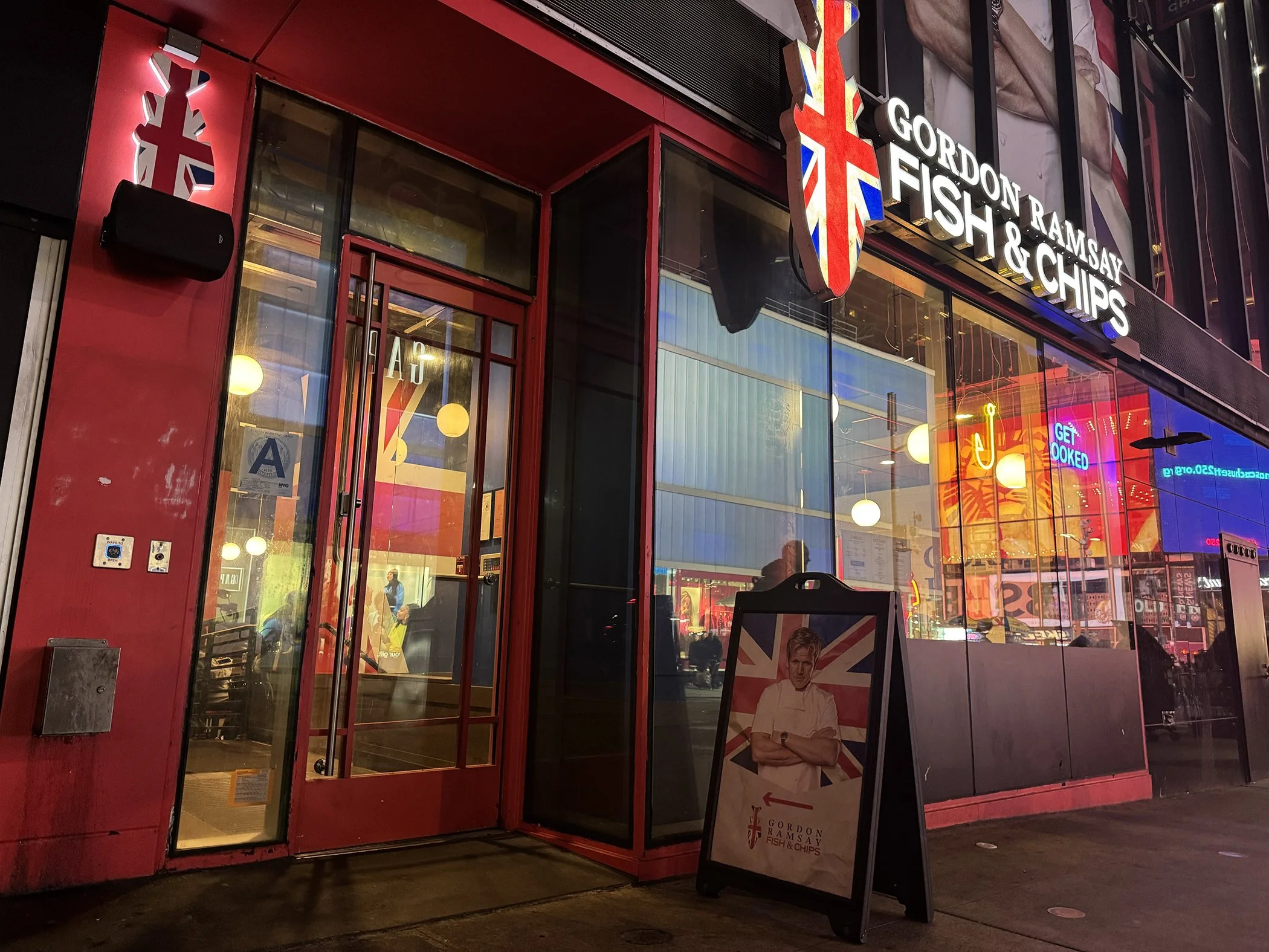 The exterior of Gordon Ramsay Fish & Chips restaurant with a Union Jack flag logo and a signboard featuring a chef in front, illuminated by colorful lights at night.