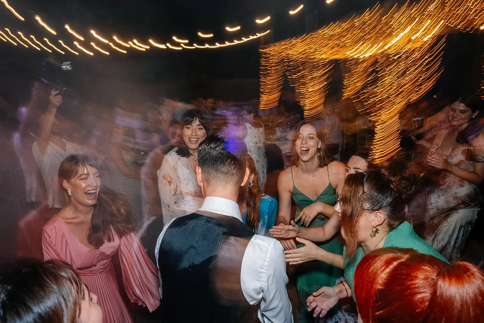 People dancing and celebrating at a party with strings of lights overhead and a lit decoration in the background.