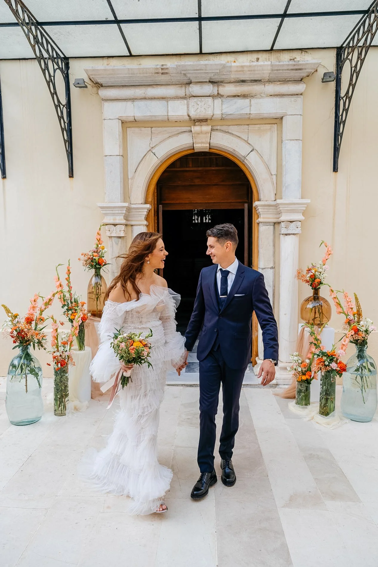 A bride and groom holding hands during their wedding ceremony, surrounded by floral arrangements in vases, outside a building with a formal stone doorway.