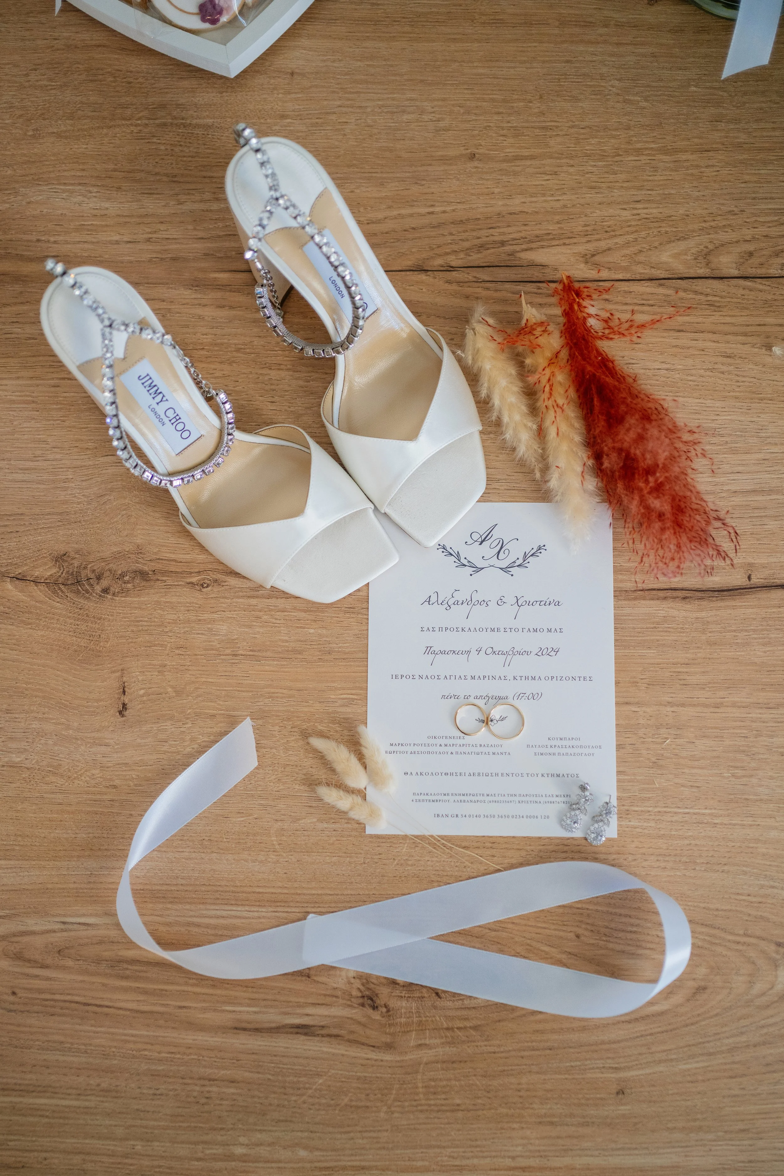 White high-heeled shoes with rhinestone straps, a wedding invitation with Greek text, gold wedding rings, and dried decorative flowers on a wooden surface.