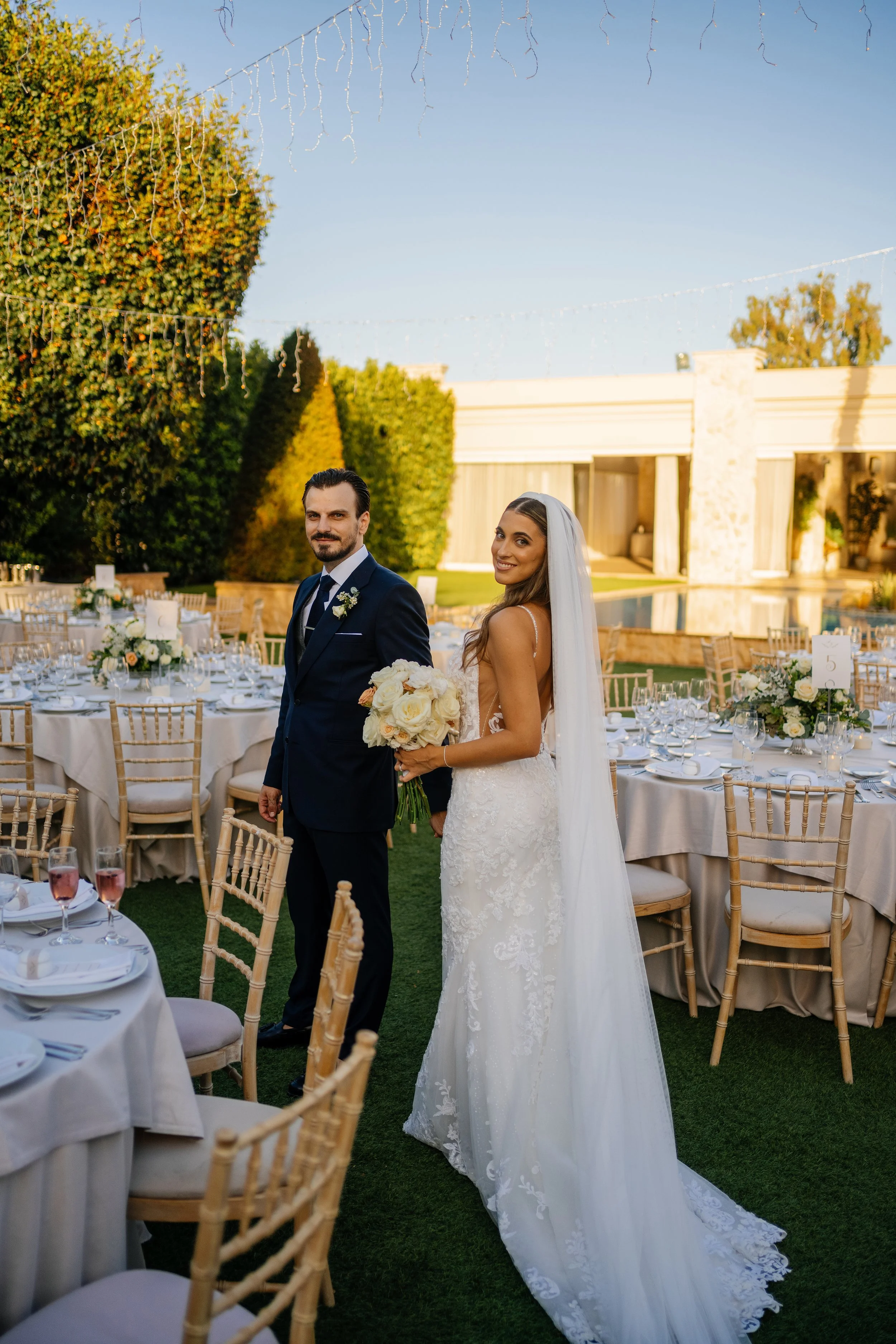 A bride and groom standing outdoors at their wedding reception, with round tables, chairs, and decorated tables in the background, under strings of fairy lights and a building with stone columns behind them.