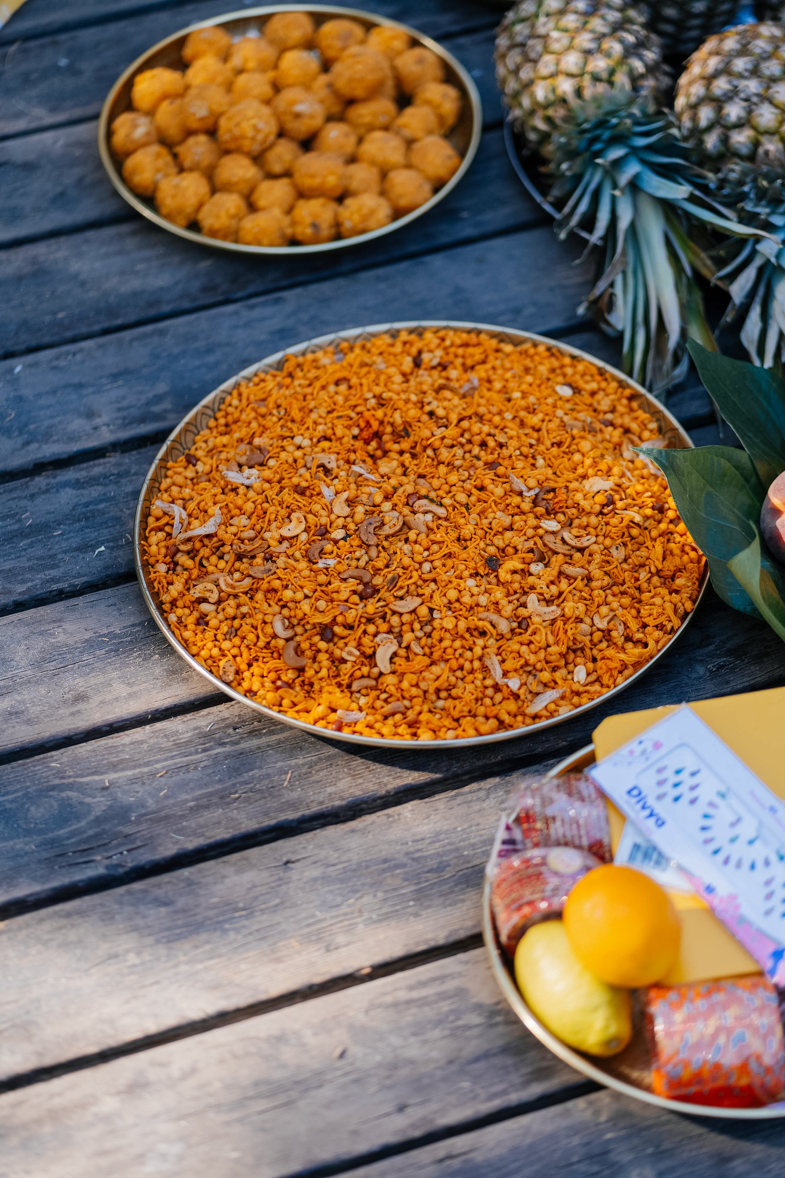 Small round orange snacks or balls in a plate, pineapples with spiky leaves, and a bowl of various yellow and red packaged candies on a wooden table.