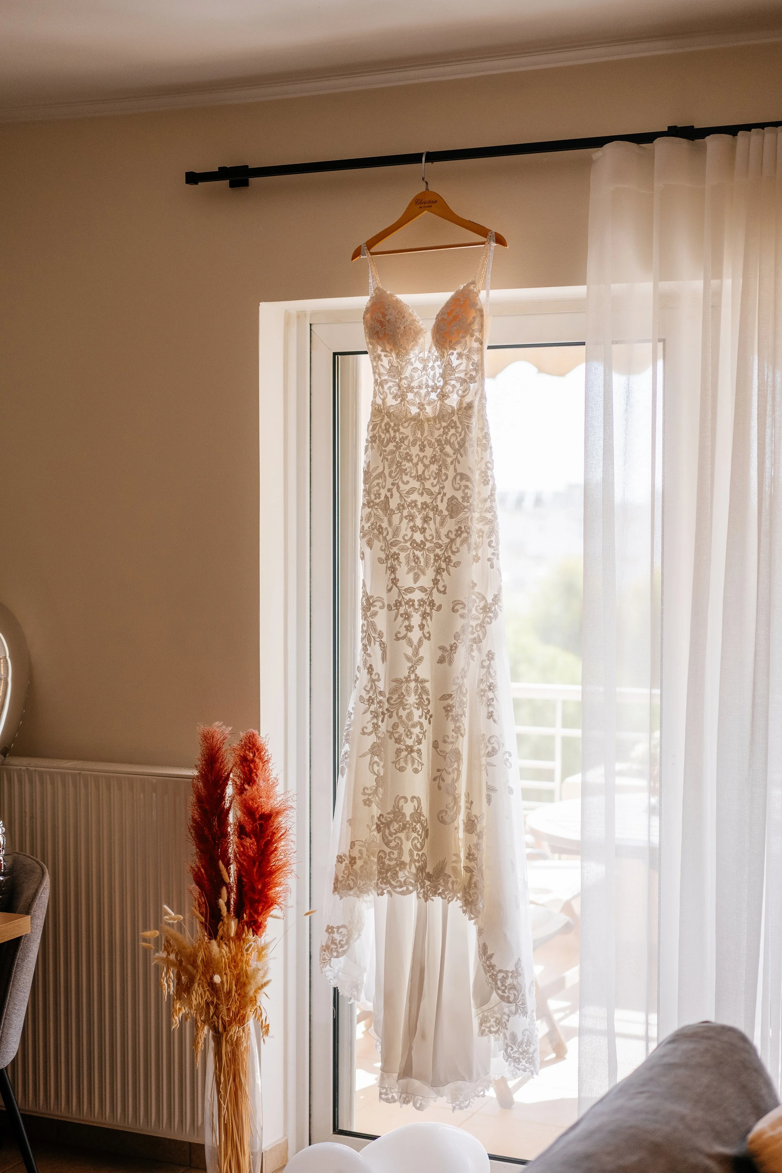 A white lace wedding dress hanging on a wooden hanger inside a room by a glass door, with sunlight coming through sheer curtains.