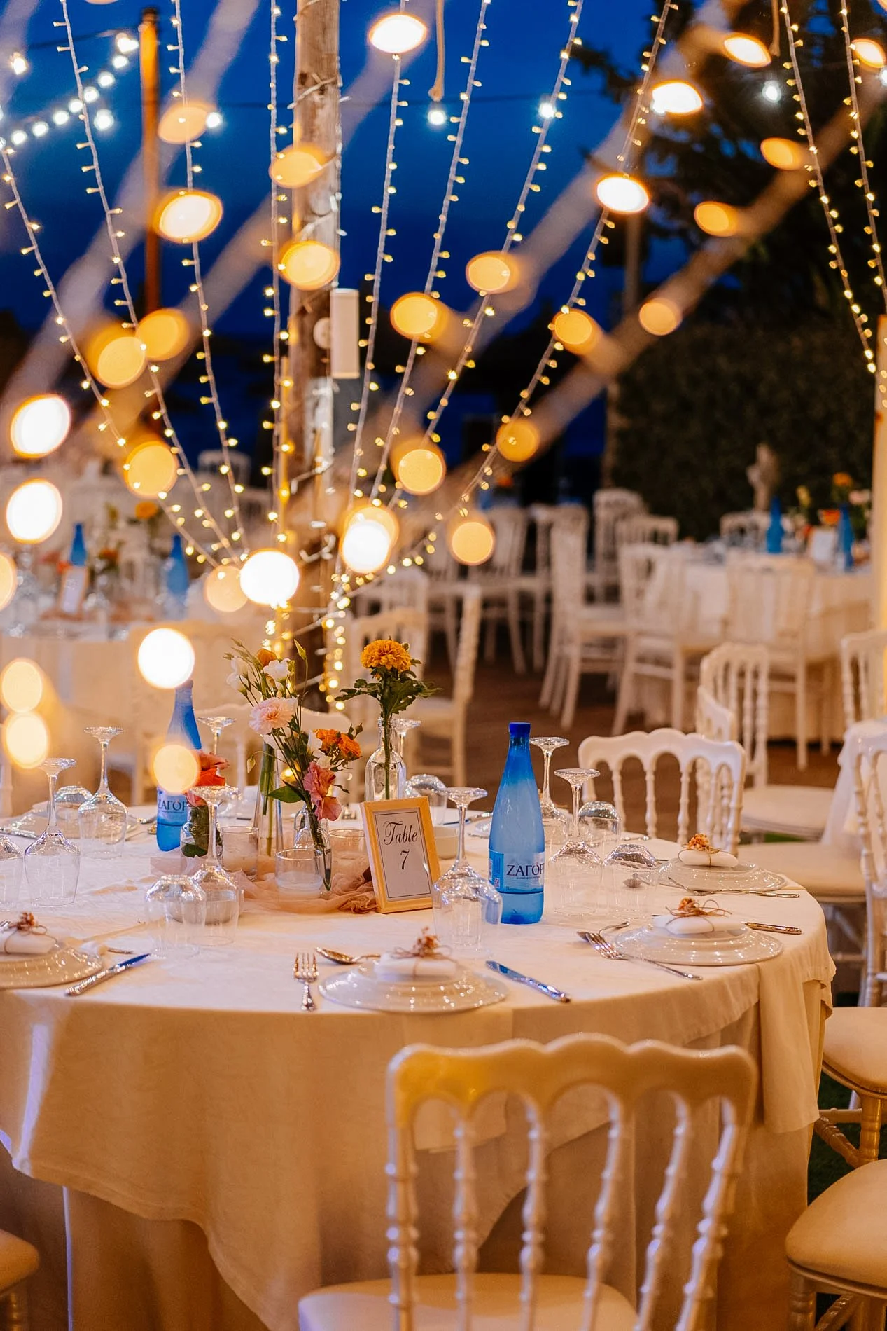 Decorated outdoor event space with string lights hanging from trees, a round table set with floral centerpieces, blue water bottles, and place settings, during evening time.