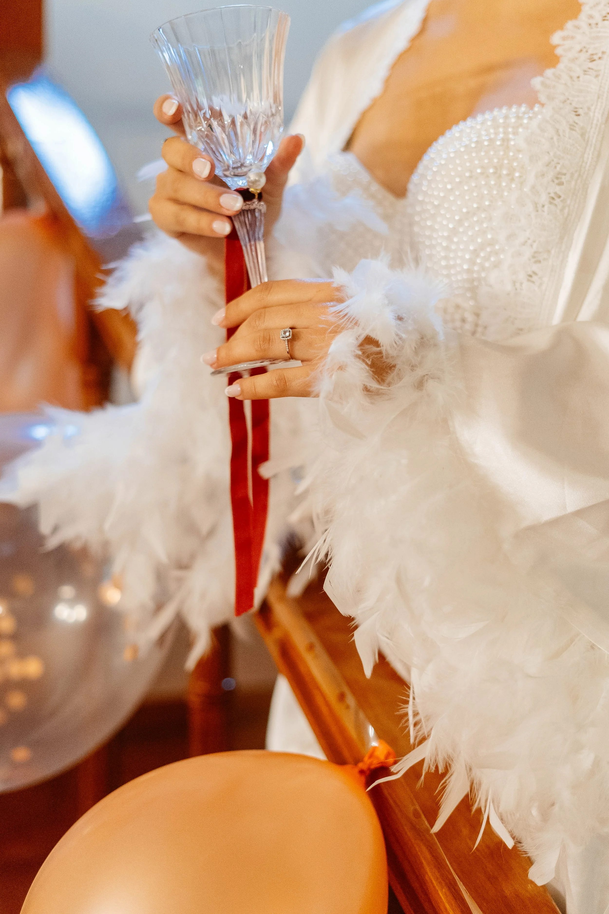 Close-up of a woman holding a glass of champagne, wearing a white feathered dress with pearl embellishments on the chest. She has a ring with a large gemstone and a pearl bracelet, and her nails are painted white. The setting appears to be indoors, p