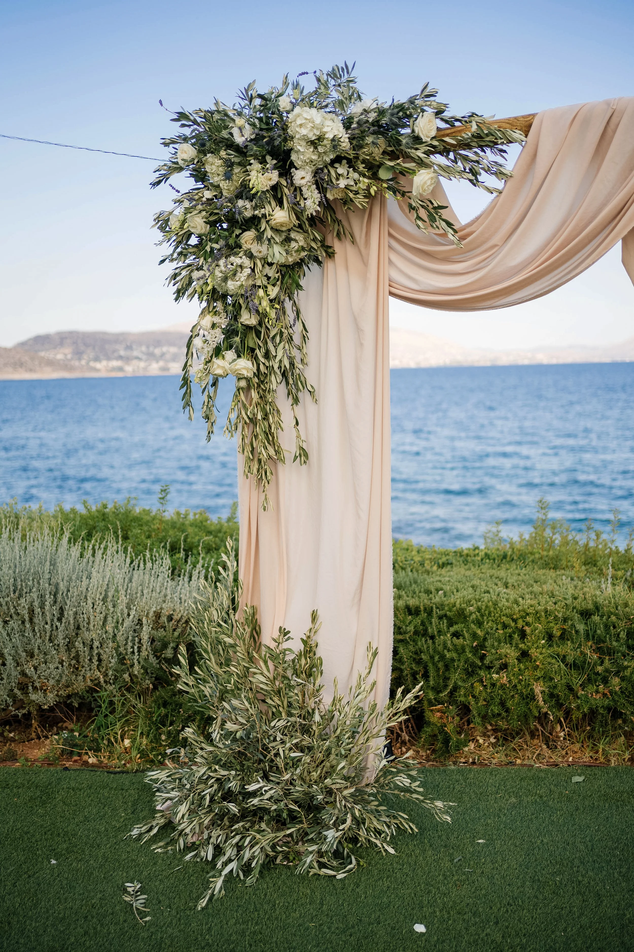 Decorative wooden arch with beige fabric draping and white floral arrangement, set outdoors by the water with hills in the background.