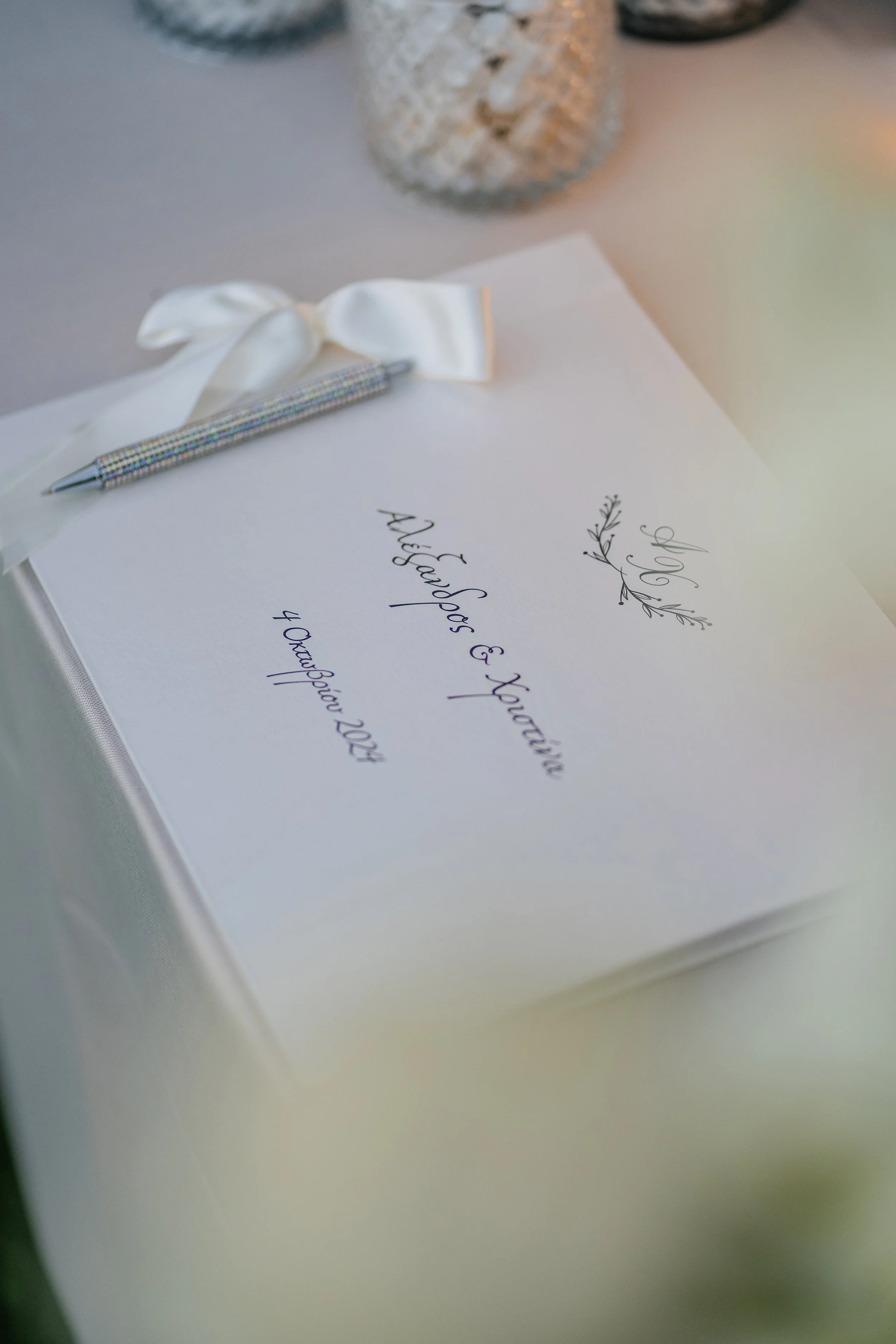 Wedding invitation card with calligraphy text and a ribbon-tied pen on top, placed on a white surface with decorative vases in the background.