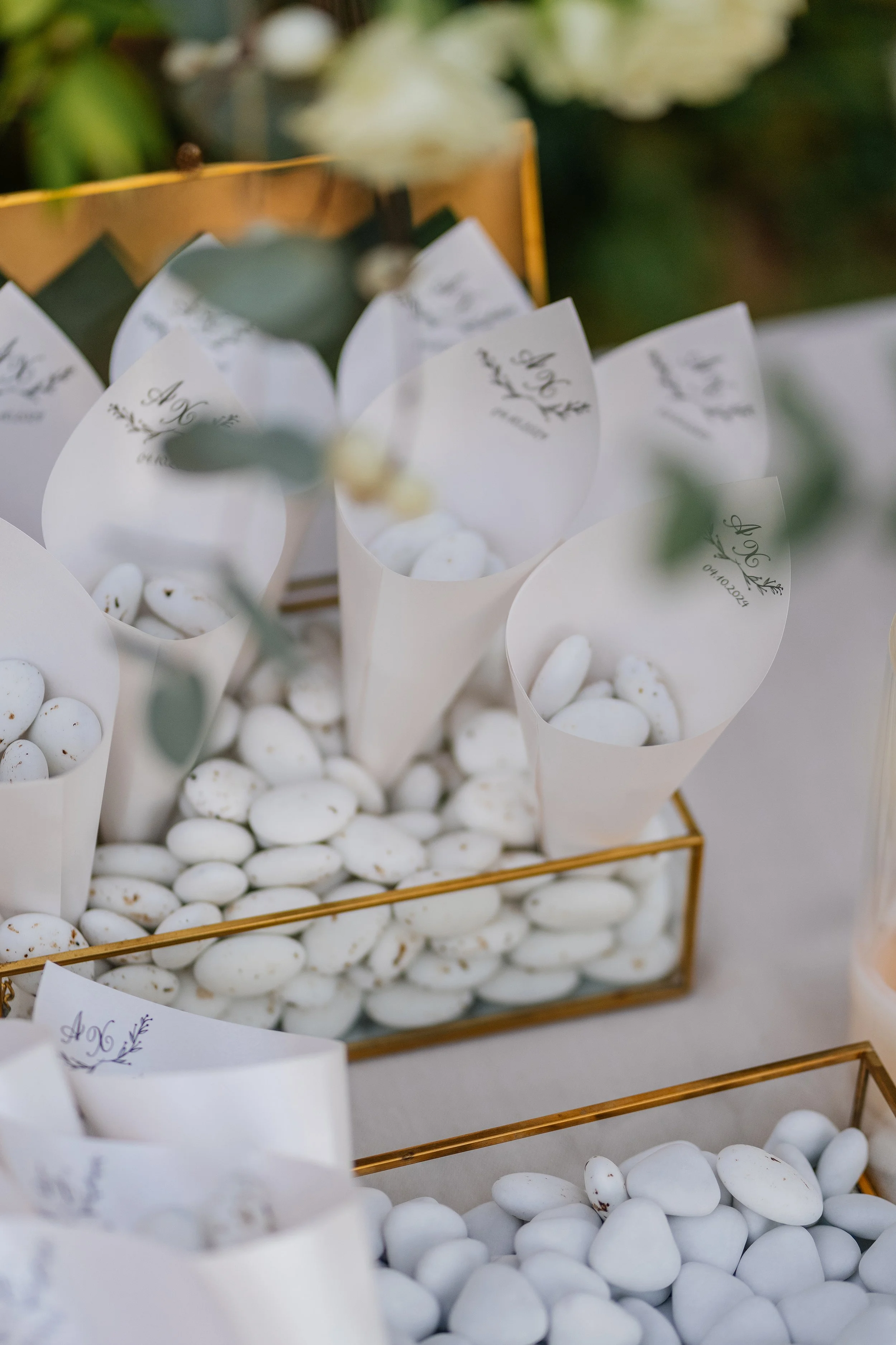 White candies in paper cones with writing, arranged in a gold wire basket, with a blurred background of white flowers and greenery.