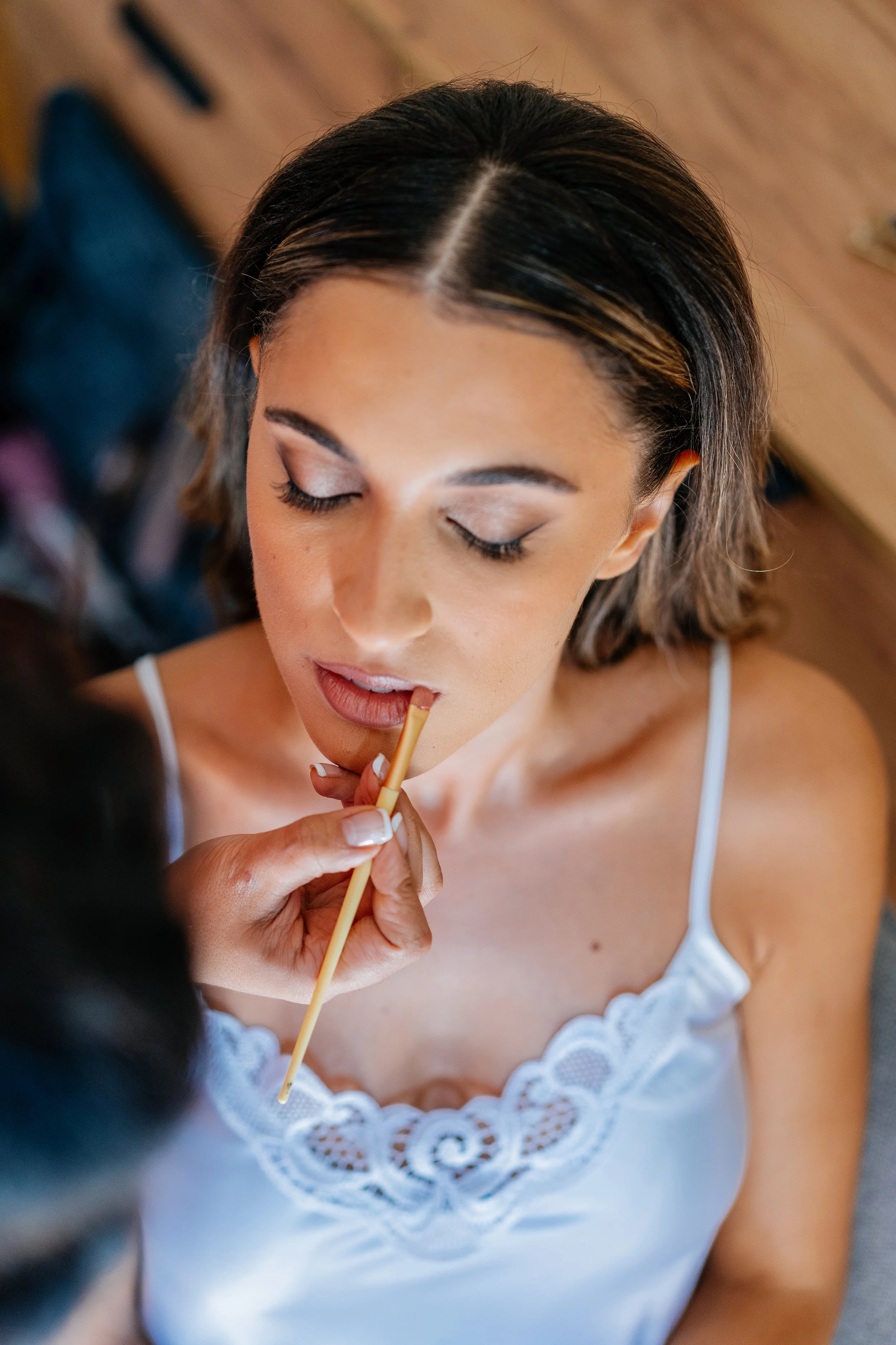 A woman with medium-length hair and a white top is having her makeup touched up.