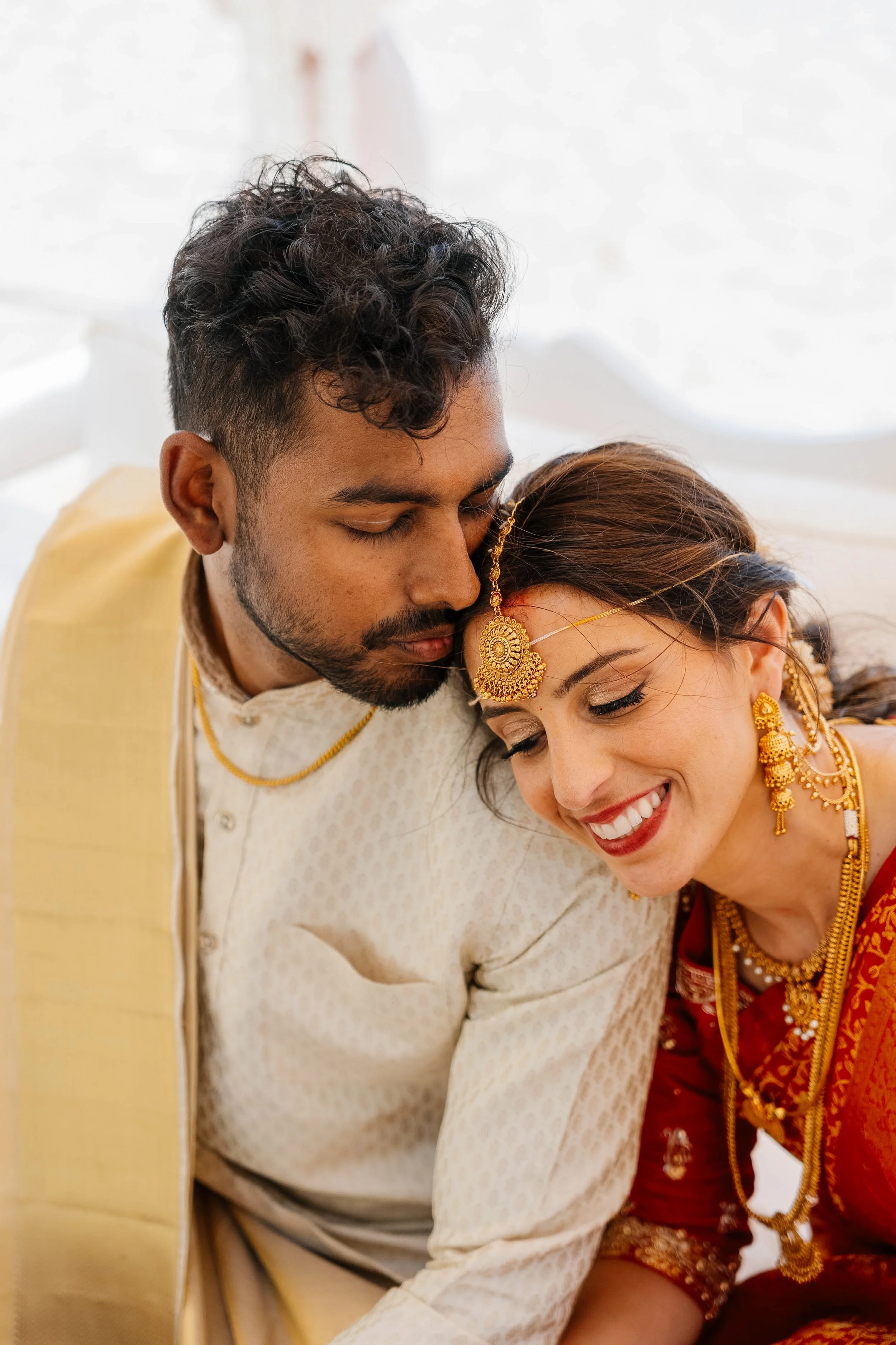 A newlywed couple, dressed in traditional Indian wedding attire, sharing an intimate moment with their foreheads touching.
