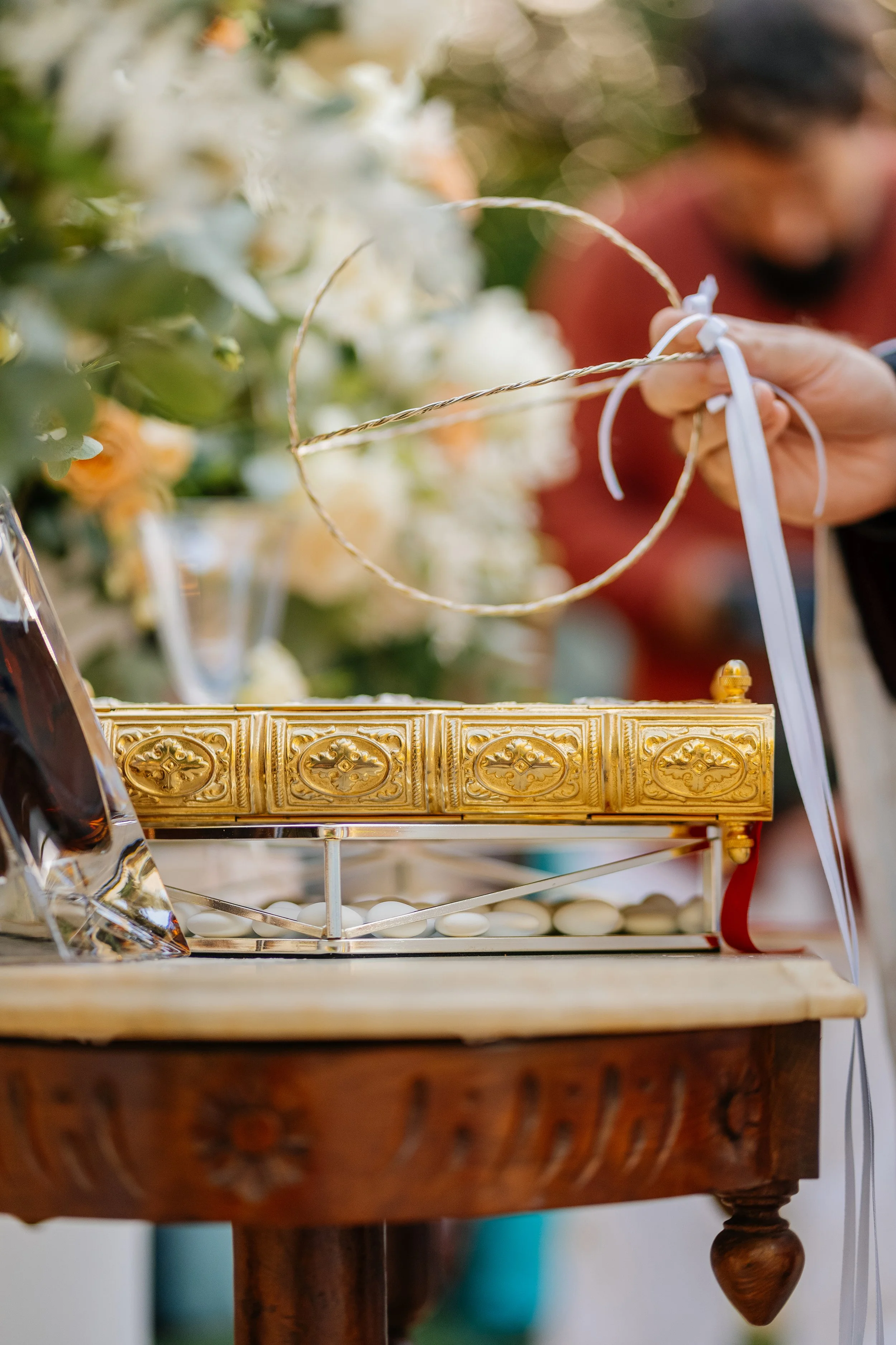 Close-up of a hand holding a white ribbon tied to a gold decorative ring, with white flowers in the background and a person wearing a red shirt partially visible.