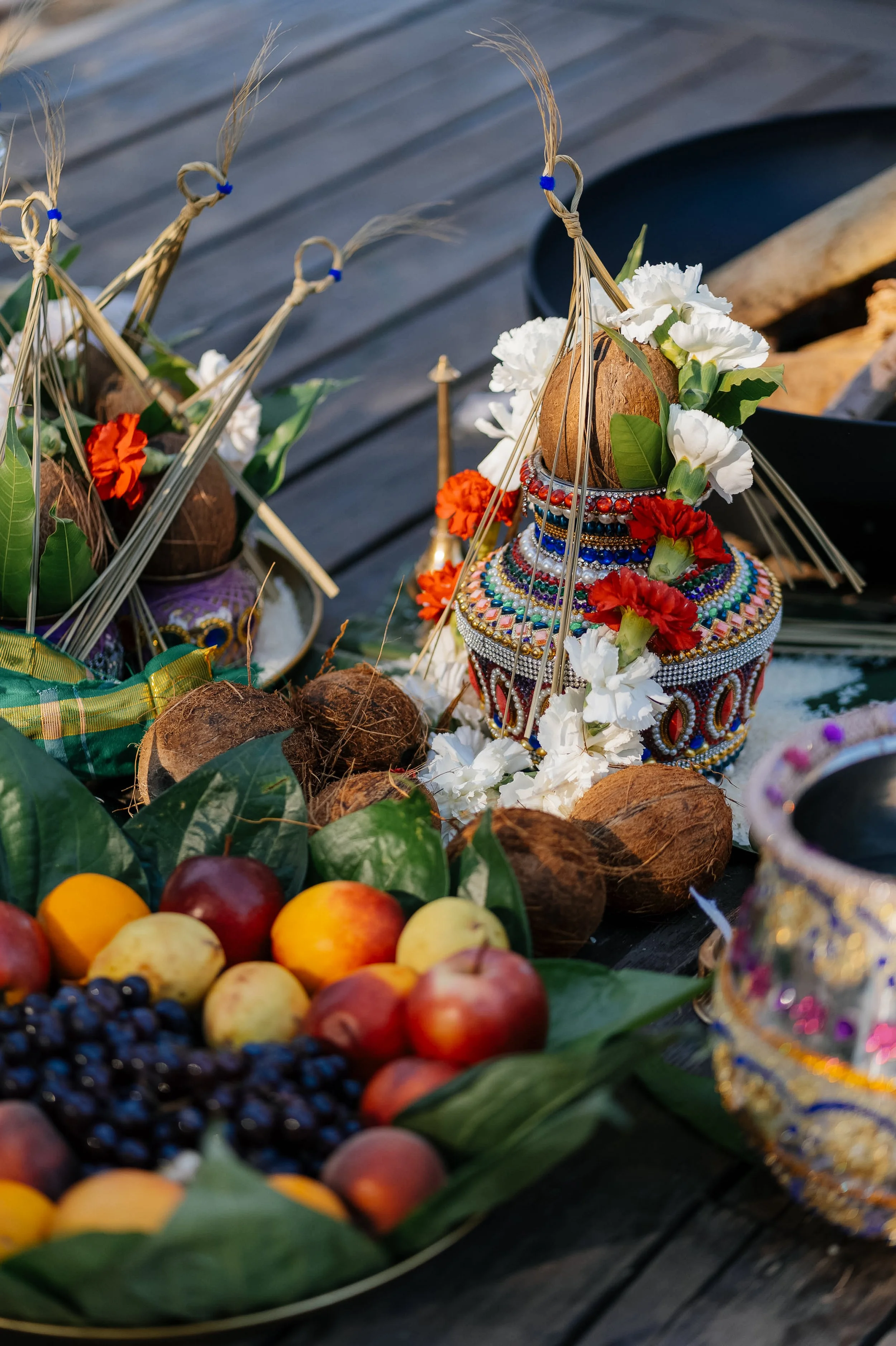 Decorative floral arrangement with white and red flowers, beads, and a coconut, surrounded by fruits like apples, plums, and blackcurrants on a wooden surface.