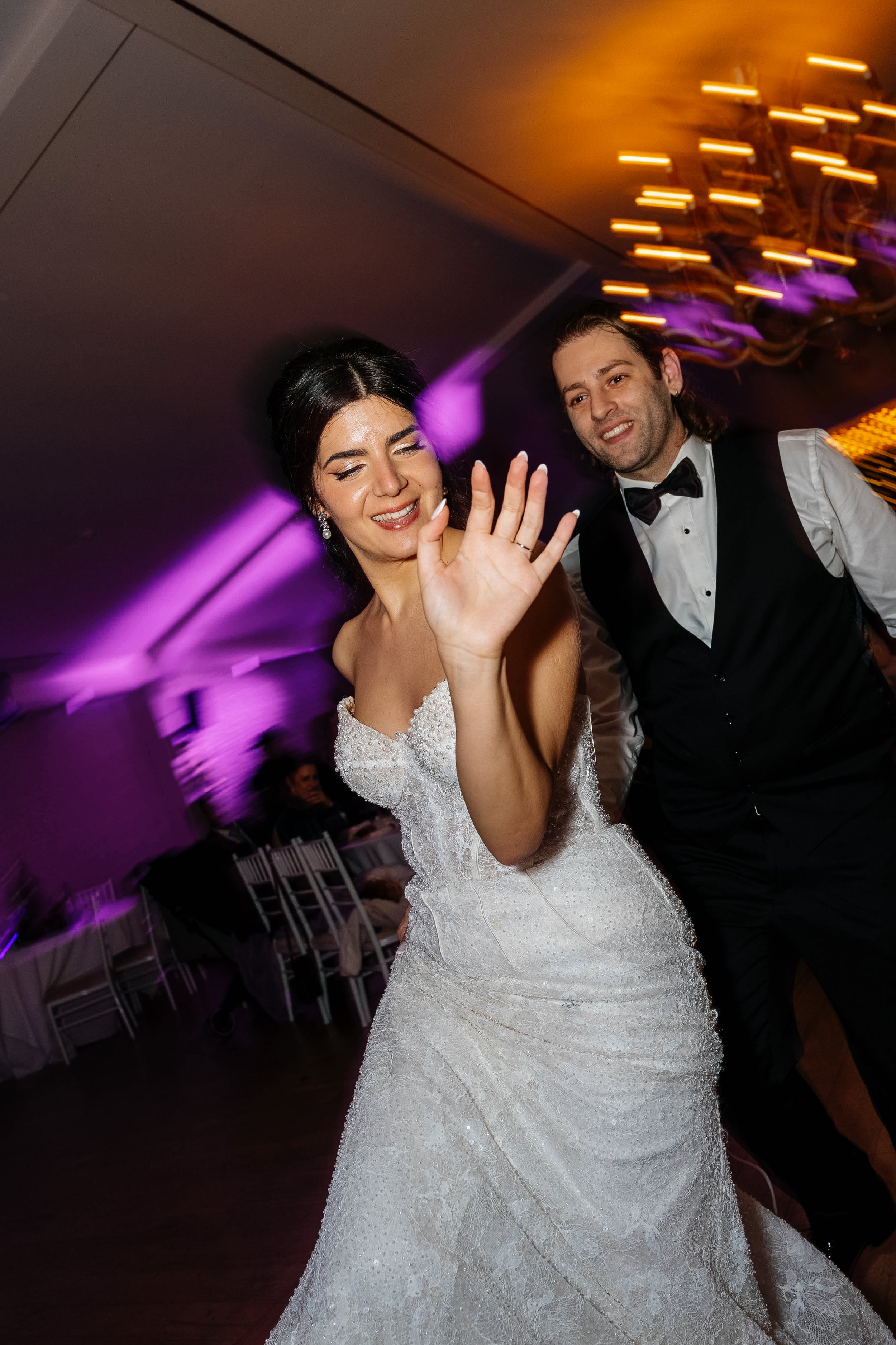 A bride in a white lacy wedding dress dancing with a groom in a black vest and bow tie at a reception, with purple and orange lighting in the background.