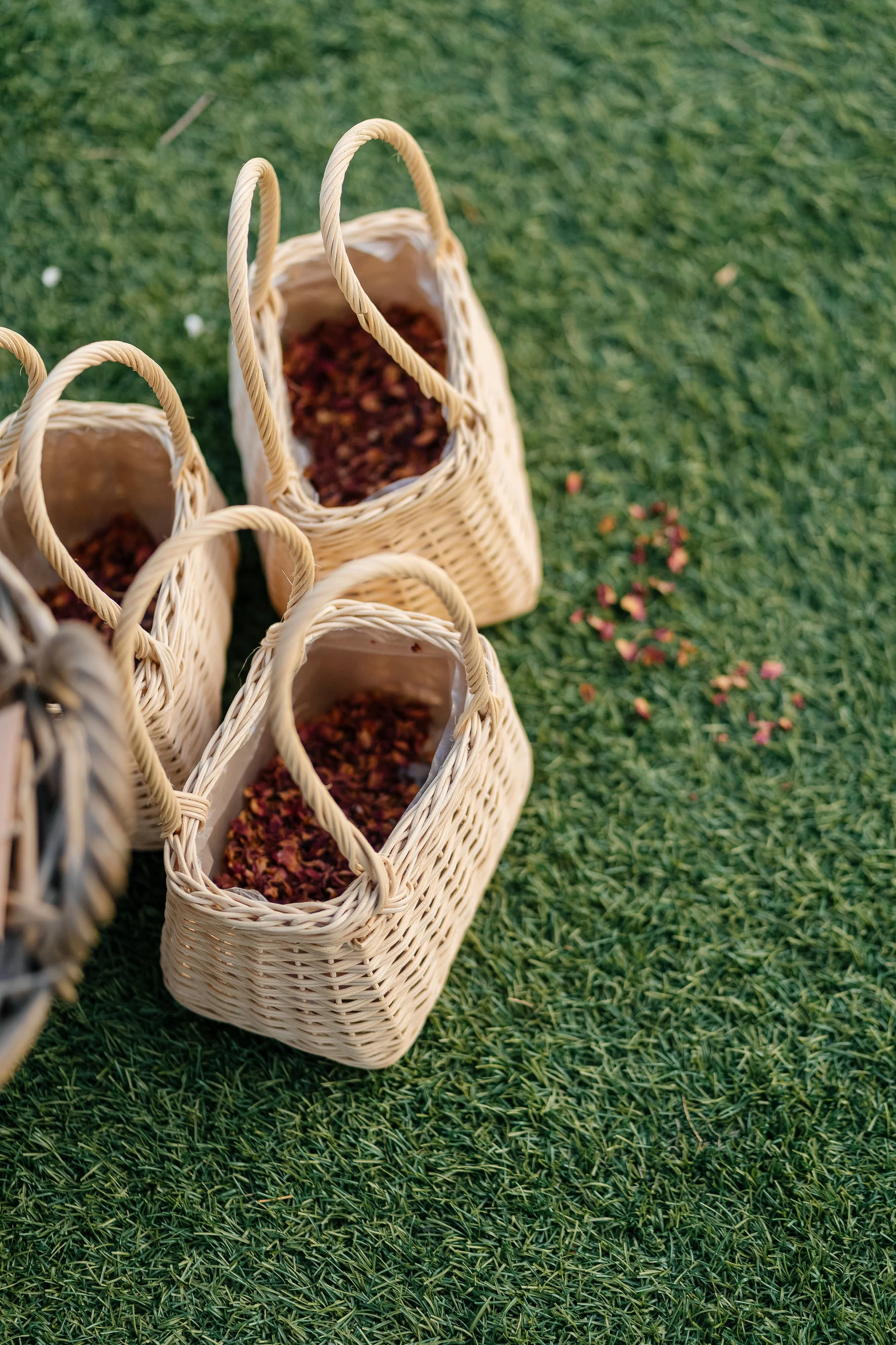 Four woven baskets filled with dried rose petals on a grassy surface.