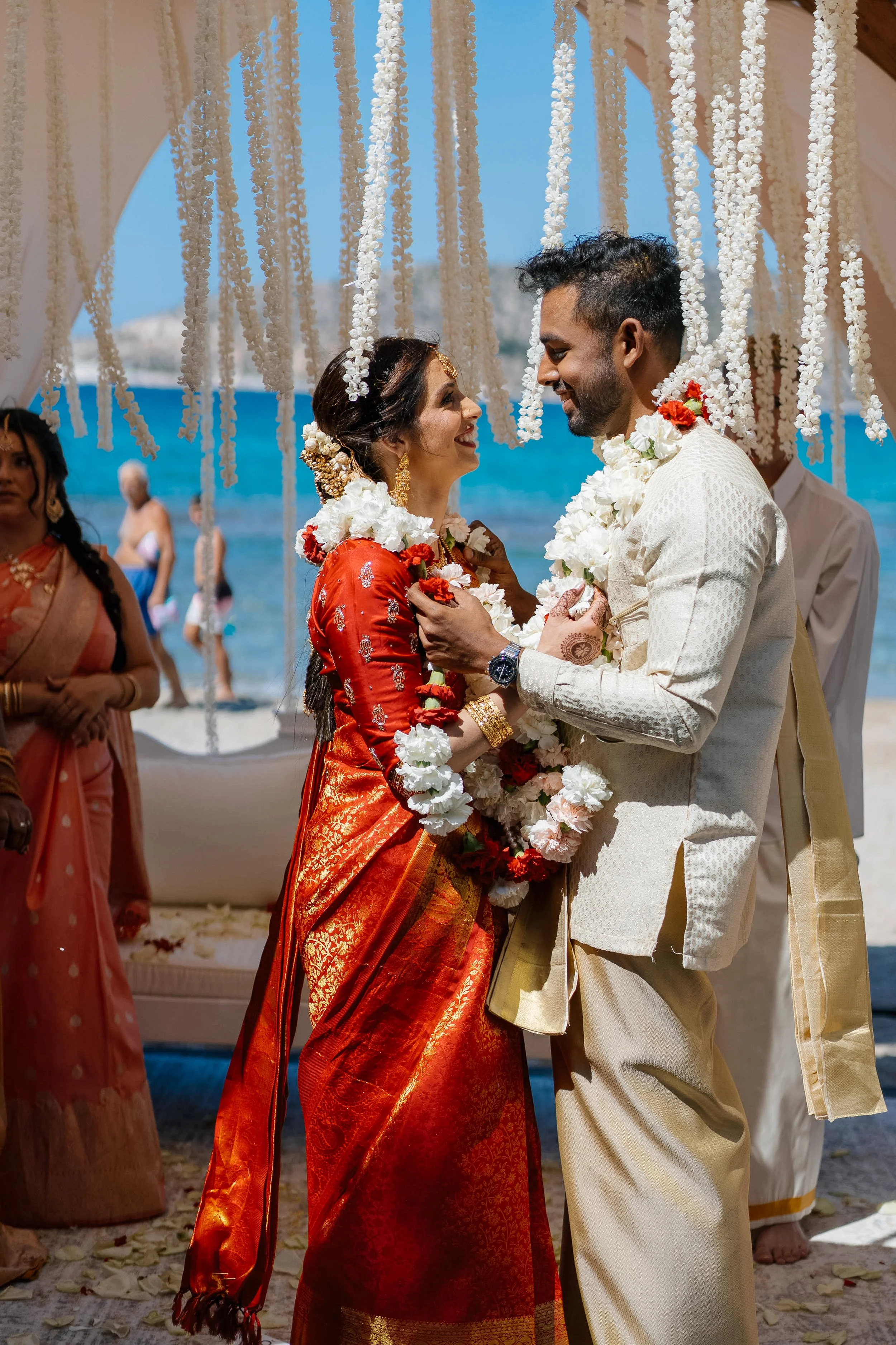 A bride and groom dressed in traditional South Asian wedding attire sharing a dance on the beach under floral decorations.