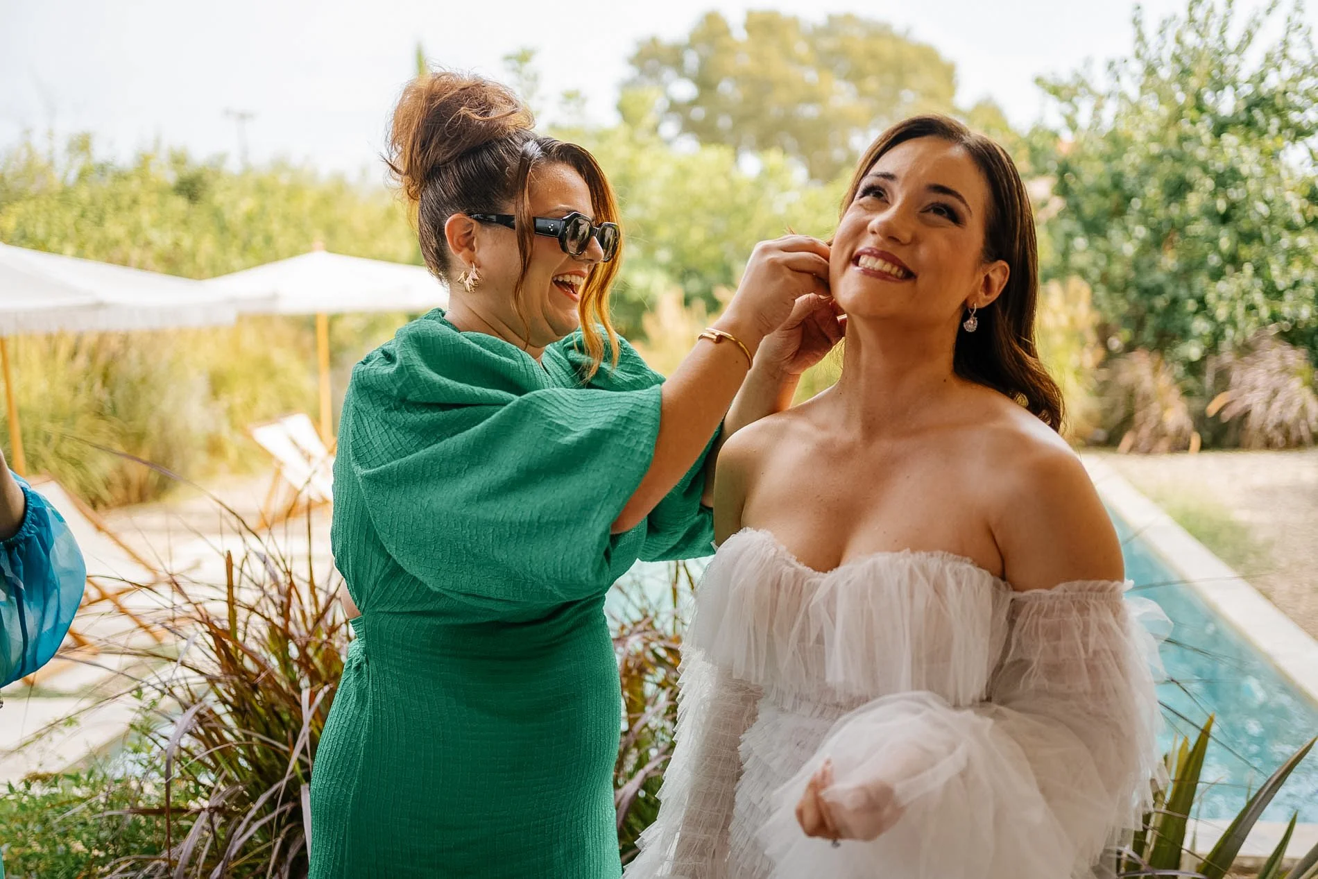 A woman in a strapless dress is smiling while another woman, wearing sunglasses and a green dress, is adjusting her earring outdoors on a sunny day.