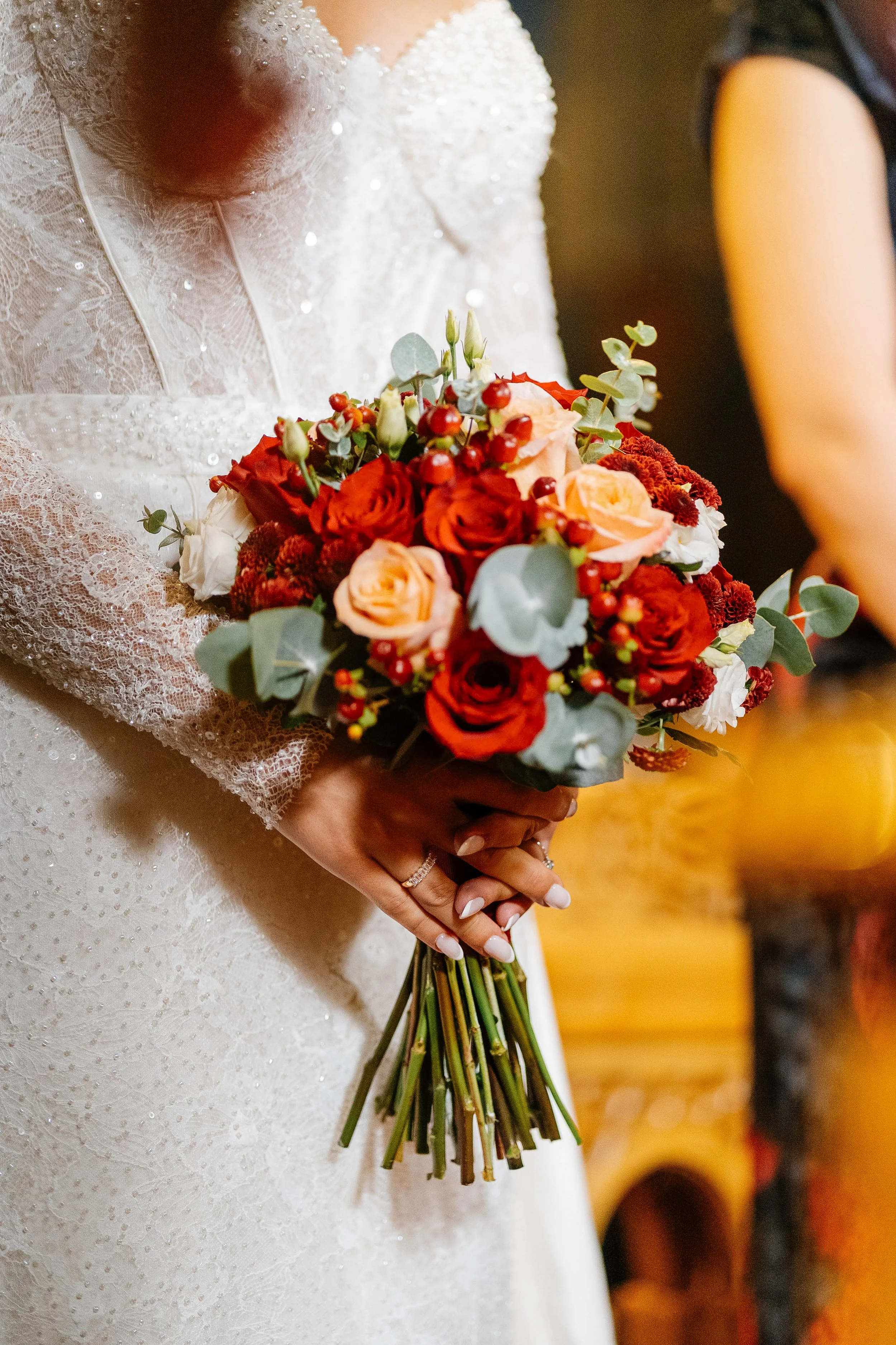 A bride dressed in a white wedding gown holding a bouquet of red, peach, and white flowers with greenery. The gown has lace and pearl embellishments. She has manicured nails and a wedding band.