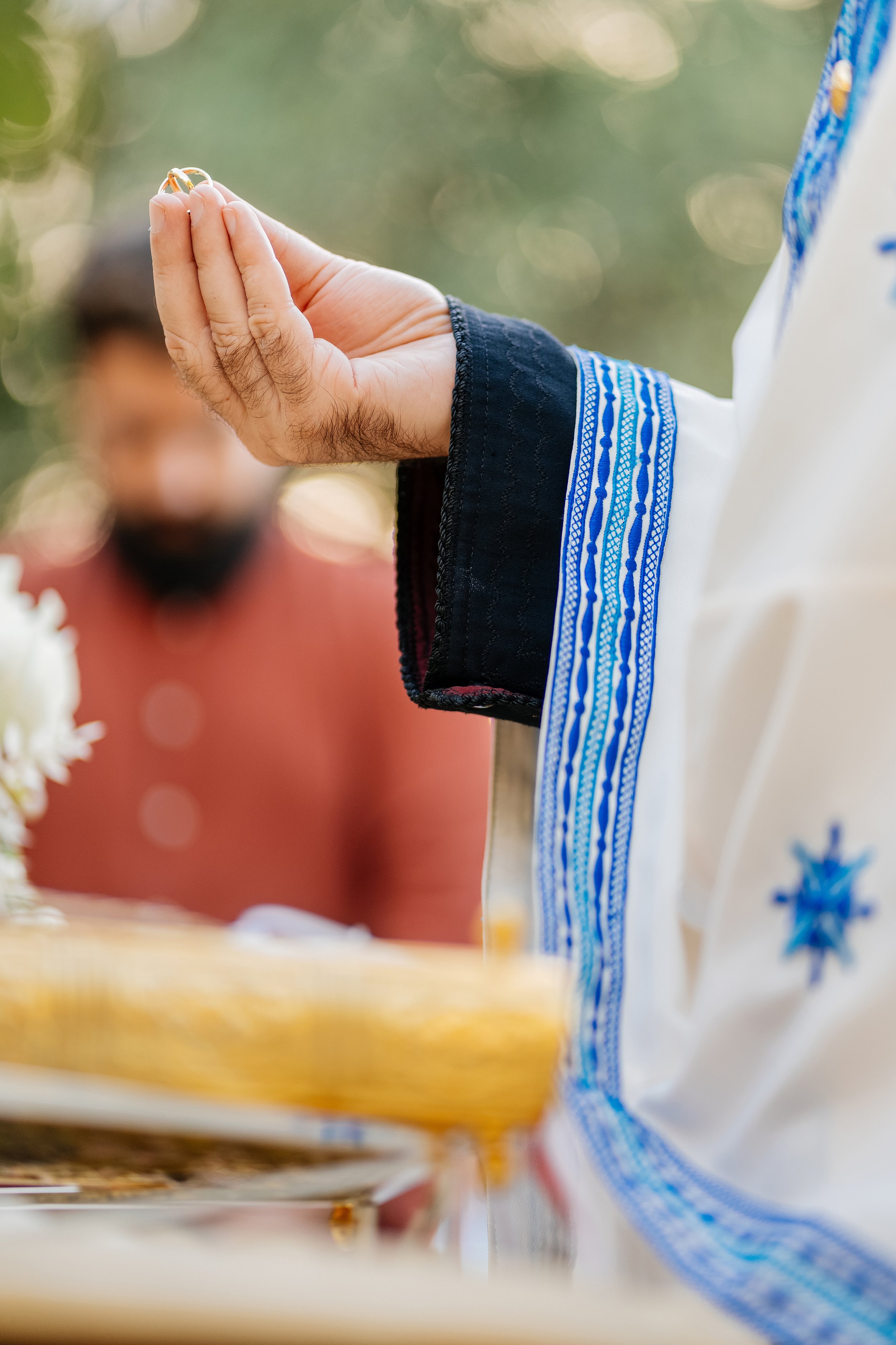Person wearing a white shirt with blue embroidery, holding a gold ring between fingers, with blurred background.