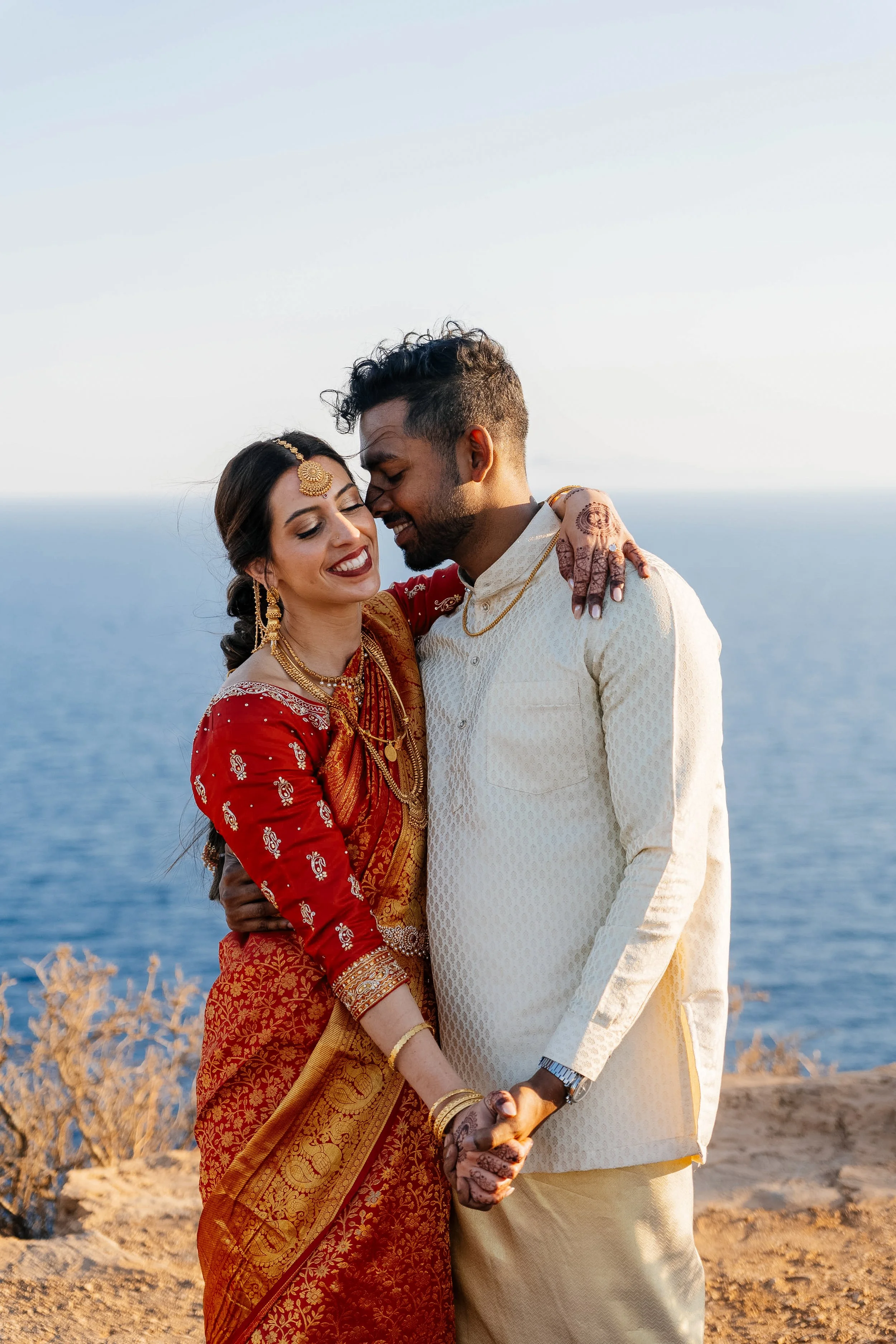 A couple dressed in traditional Indian wedding attire, holding hands, standing close to each other with eyes closed and smiling, near the ocean with a clear sky in the background.