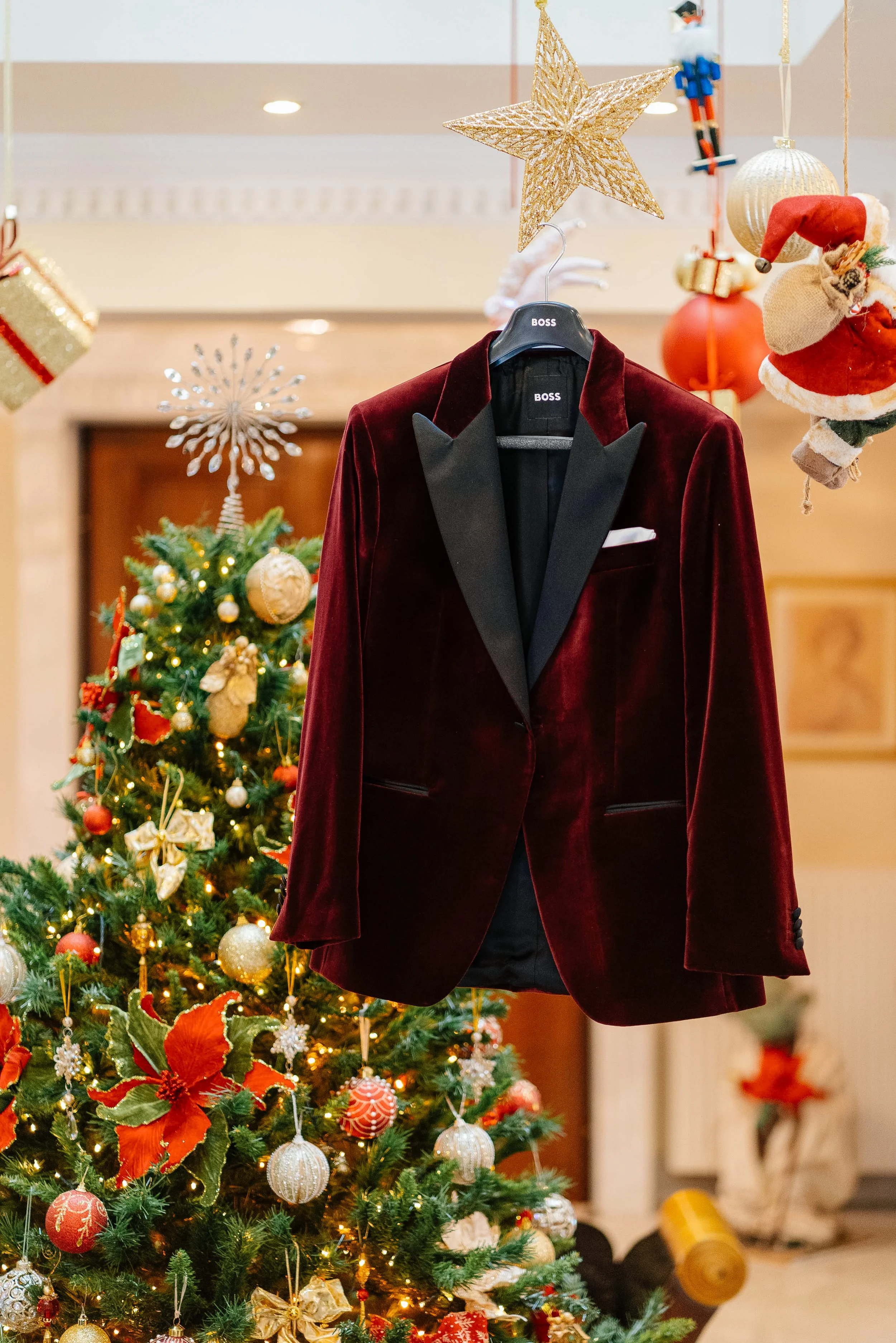 A Christmas tree decorated with gold and red ornaments, gold ribbons, and lights, standing in a room with a fire place. Above the tree hangs a gold star, and Christmas stockings, Santa hat, and holiday ornaments are visible. In the foreground, a red 