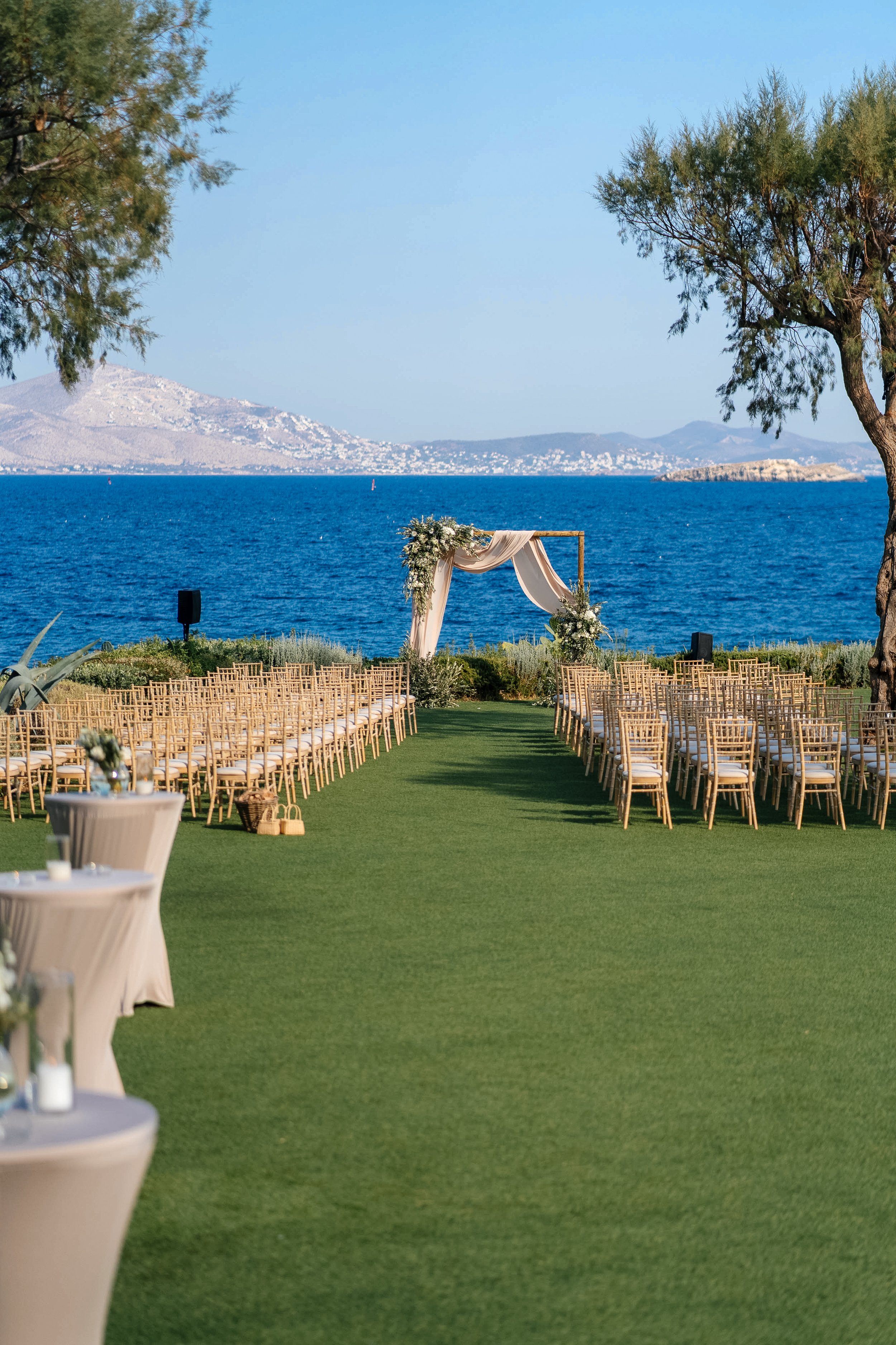 Outdoor wedding setup on a grass field by the water, with rows of chairs facing a wedding arch draped with fabric and decorated with flowers, flanked by trees under a clear blue sky.