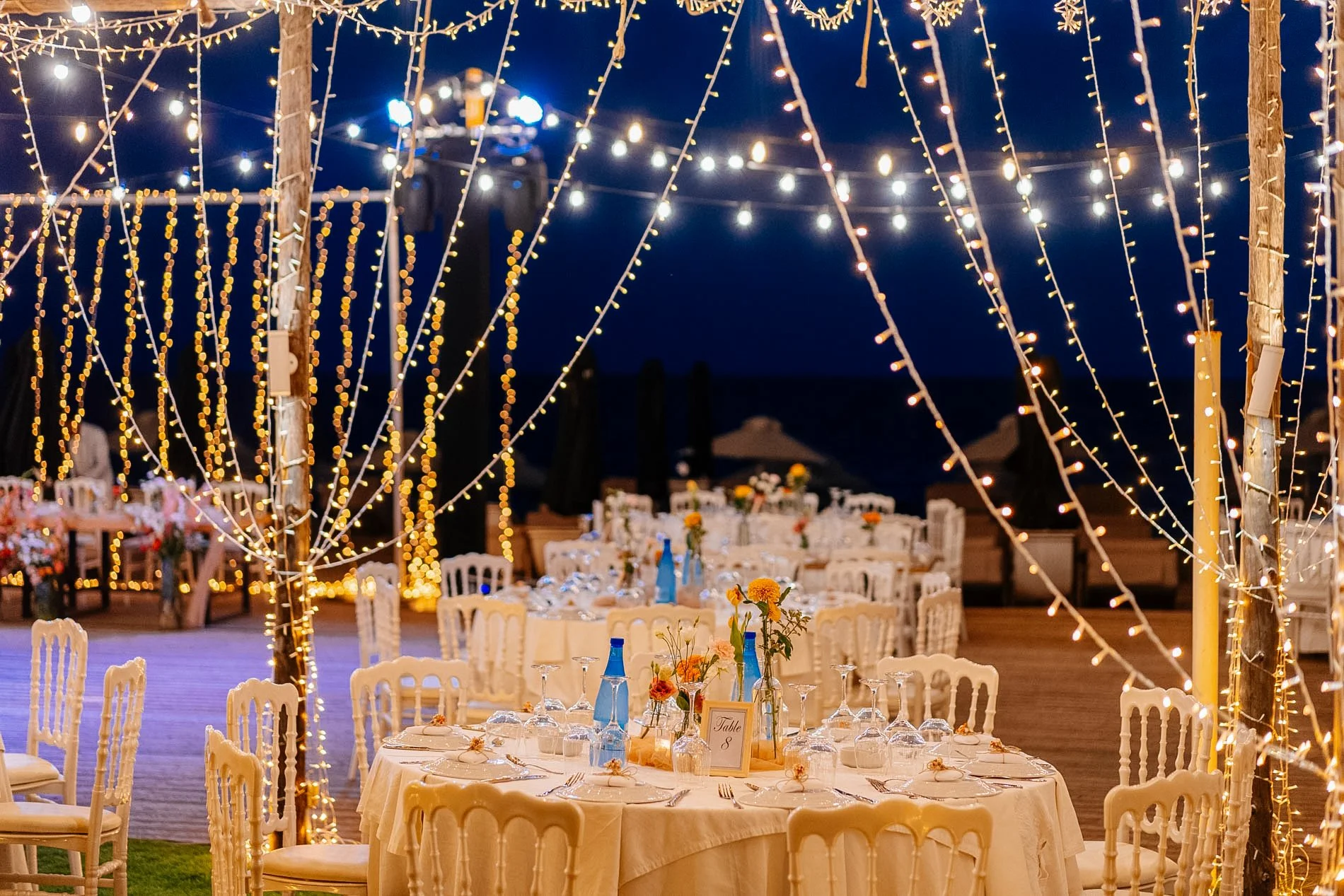 Nighttime outdoor wedding reception with string lights hanging across wooden posts, decorated tables with white chairs, floral centerpieces, and blue water bottles on tables.
