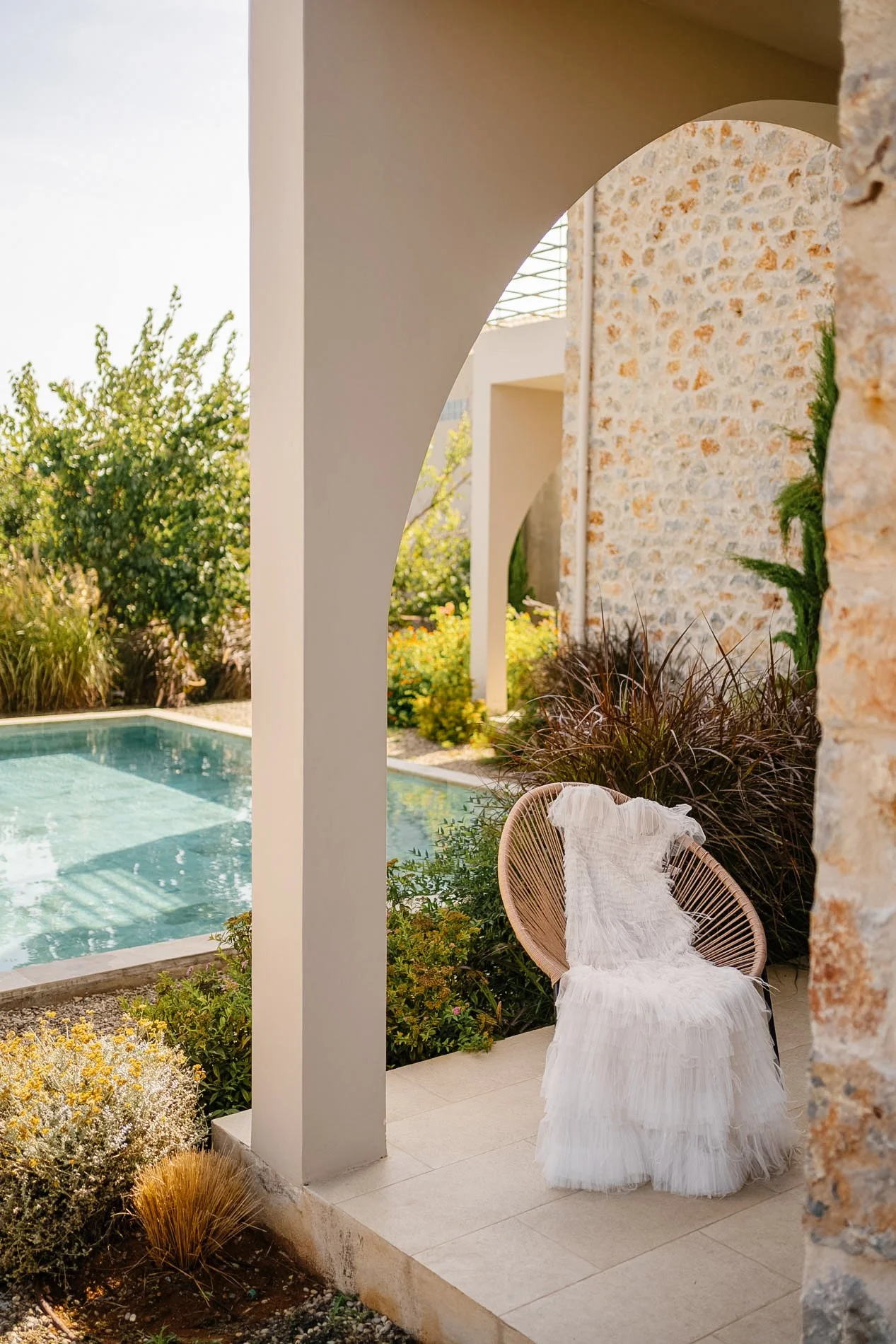 White woven chair with a pleated white fabric throw sitting on a patio with stone and stucco walls, overlooking a swimming pool surrounded by green bushes and trees.
