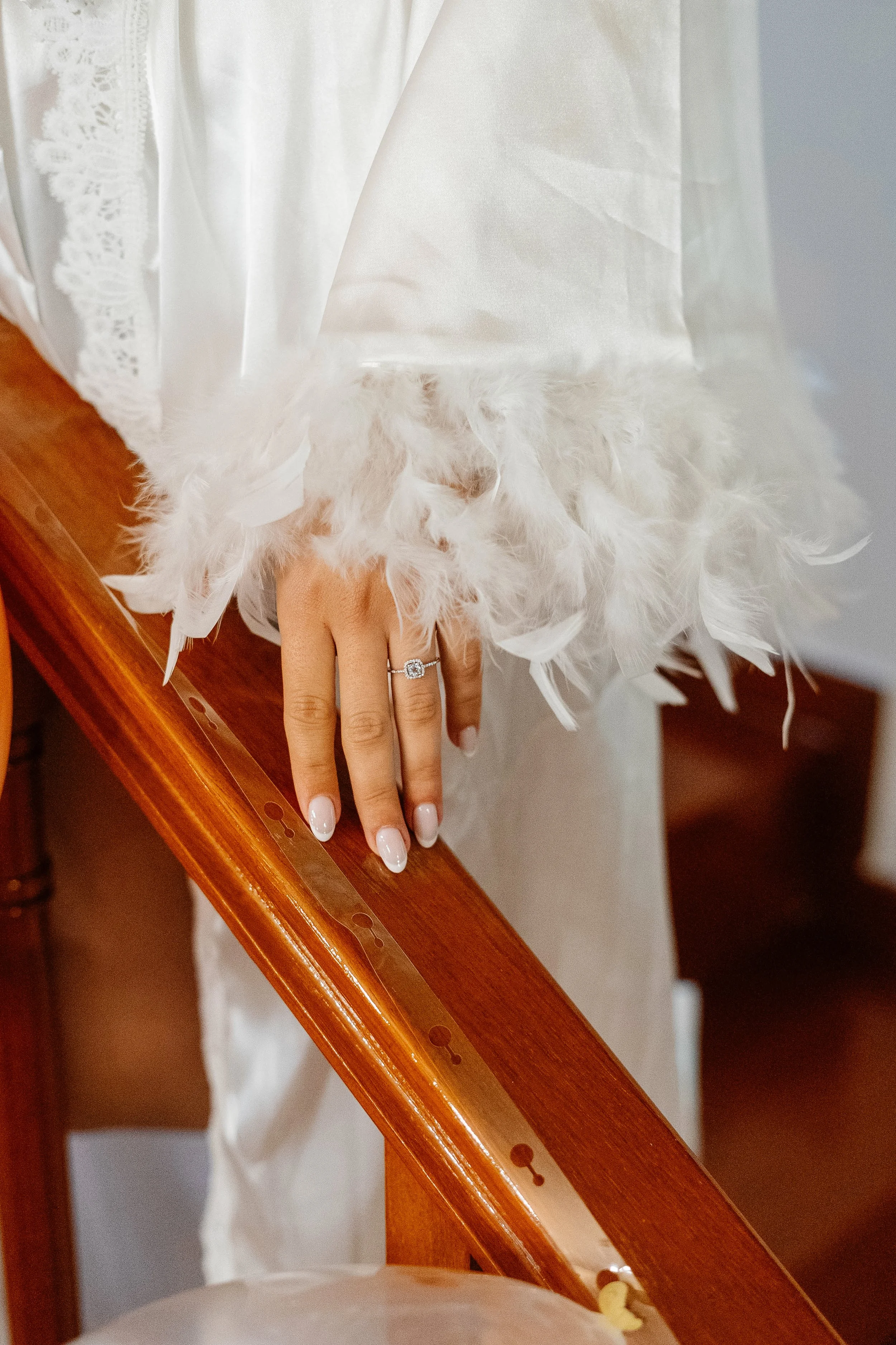 A hand with a diamond ring resting on a wooden staircase, with a white dress with feather and lace details in the background.