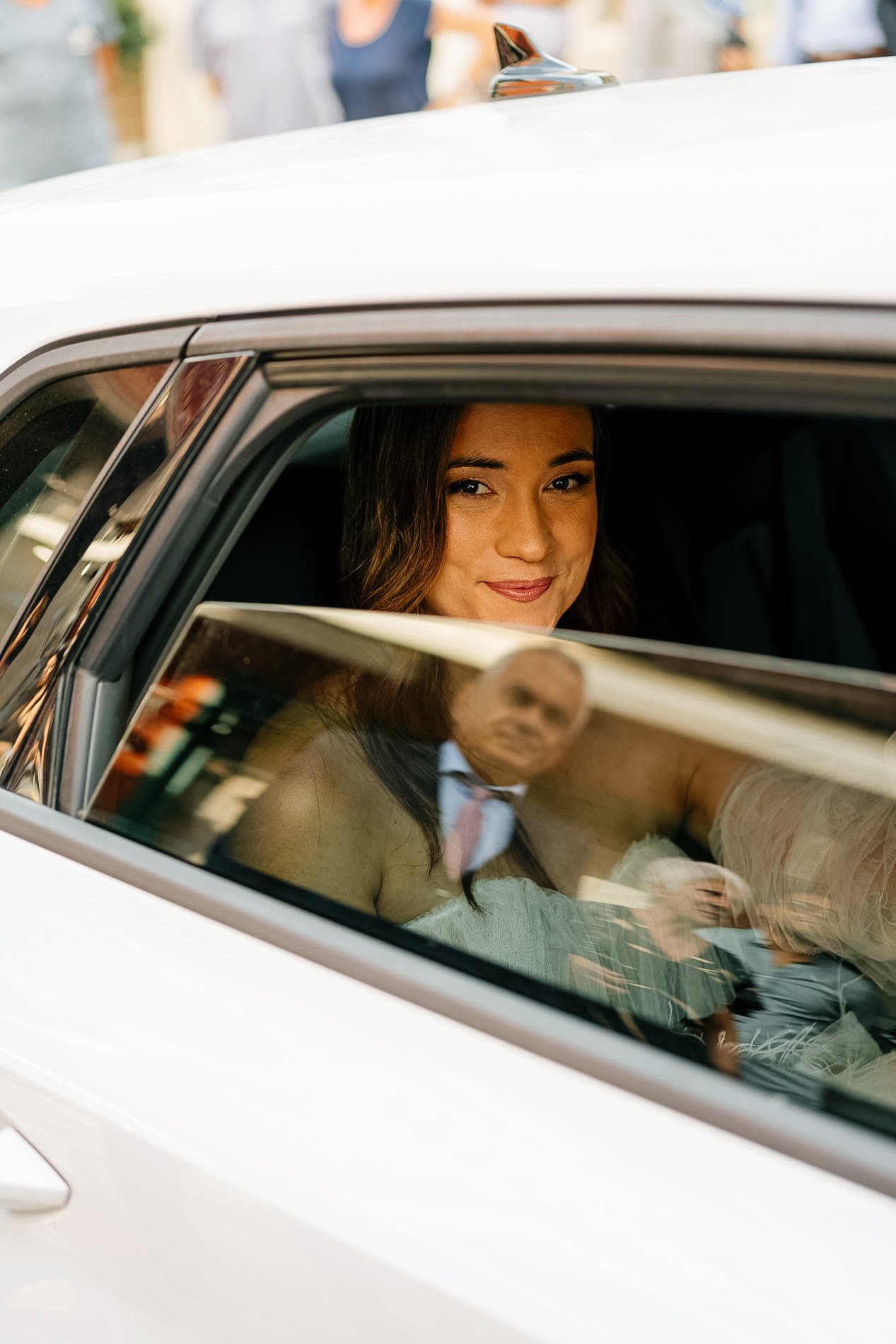 Woman smiling and looking out the car window, with an older man and woman visible inside the car. The image is focused on the woman, with a blurred background and reflections on the window.