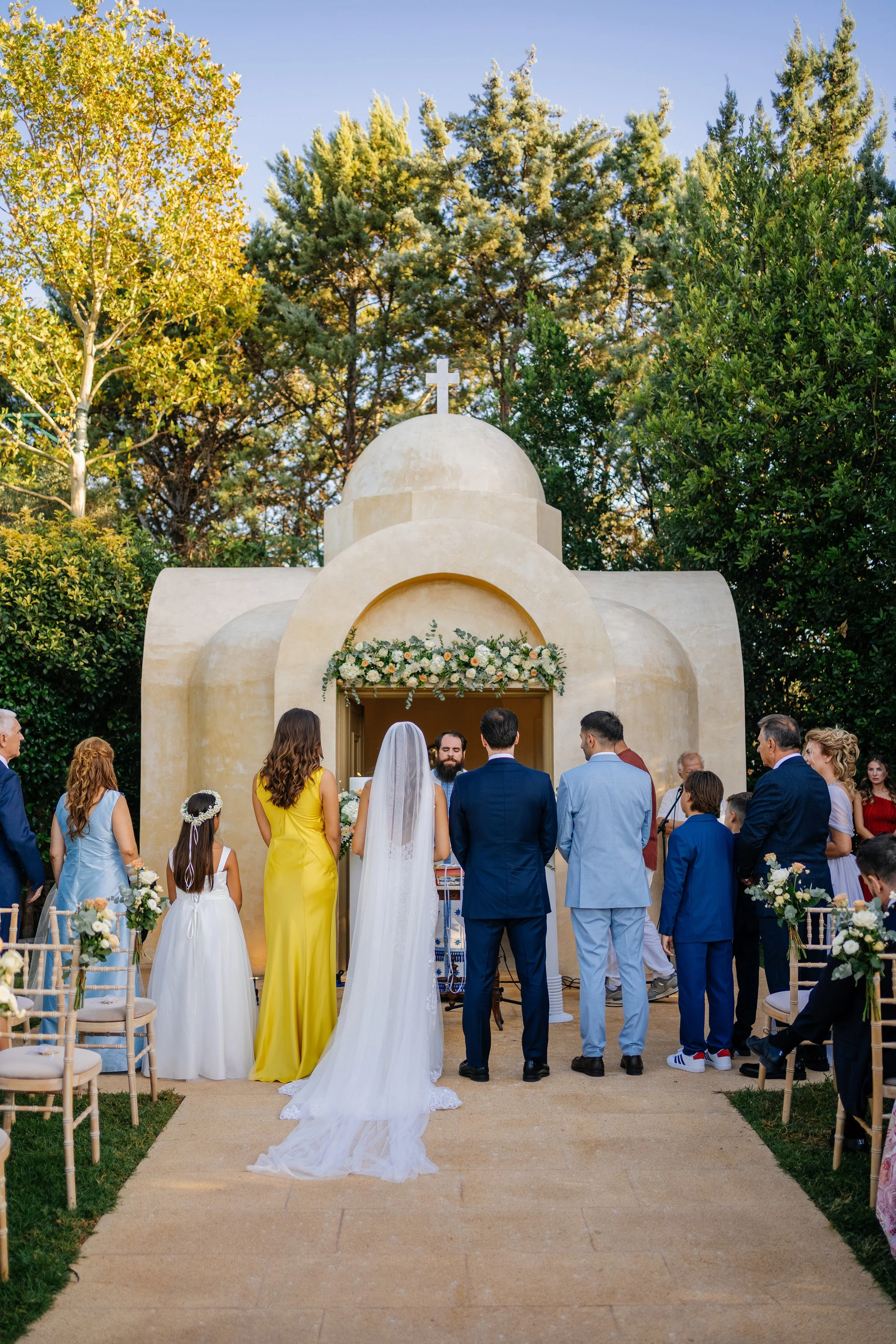 A wedding ceremony in front of a small chapel with a cross on top, decorated with flowers, and an archway. The bride in a white dress and veil stands with her back to the camera, surrounded by guests in formal attire. The scene is outdoors with trees